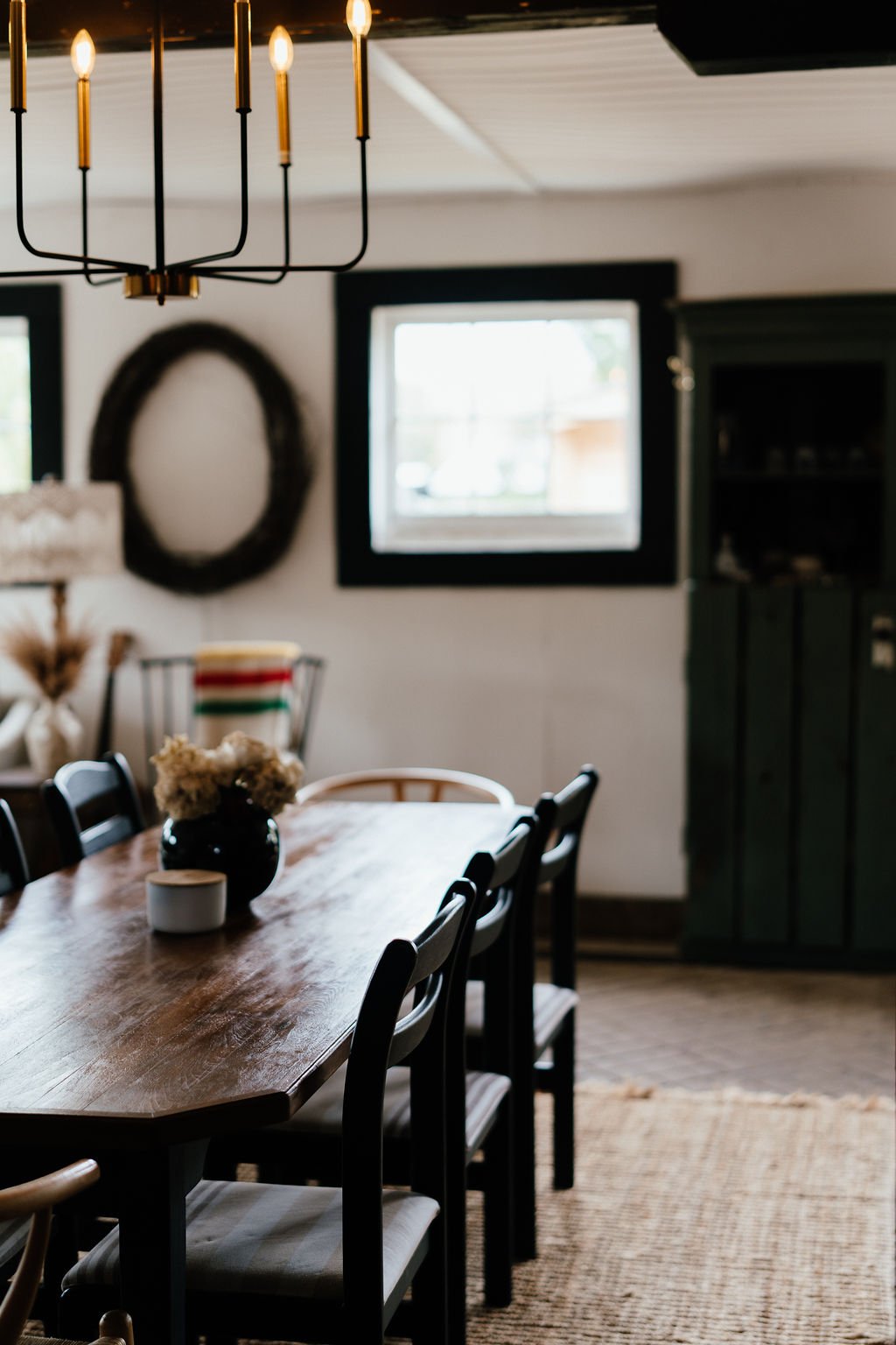 A wooden dining table with black chairs in a well-lit room. A black vase with dried flowers and a small white container are on the table. A black-framed window and a dark green cabinet are visible in the background. A modern chandelier with six lit b