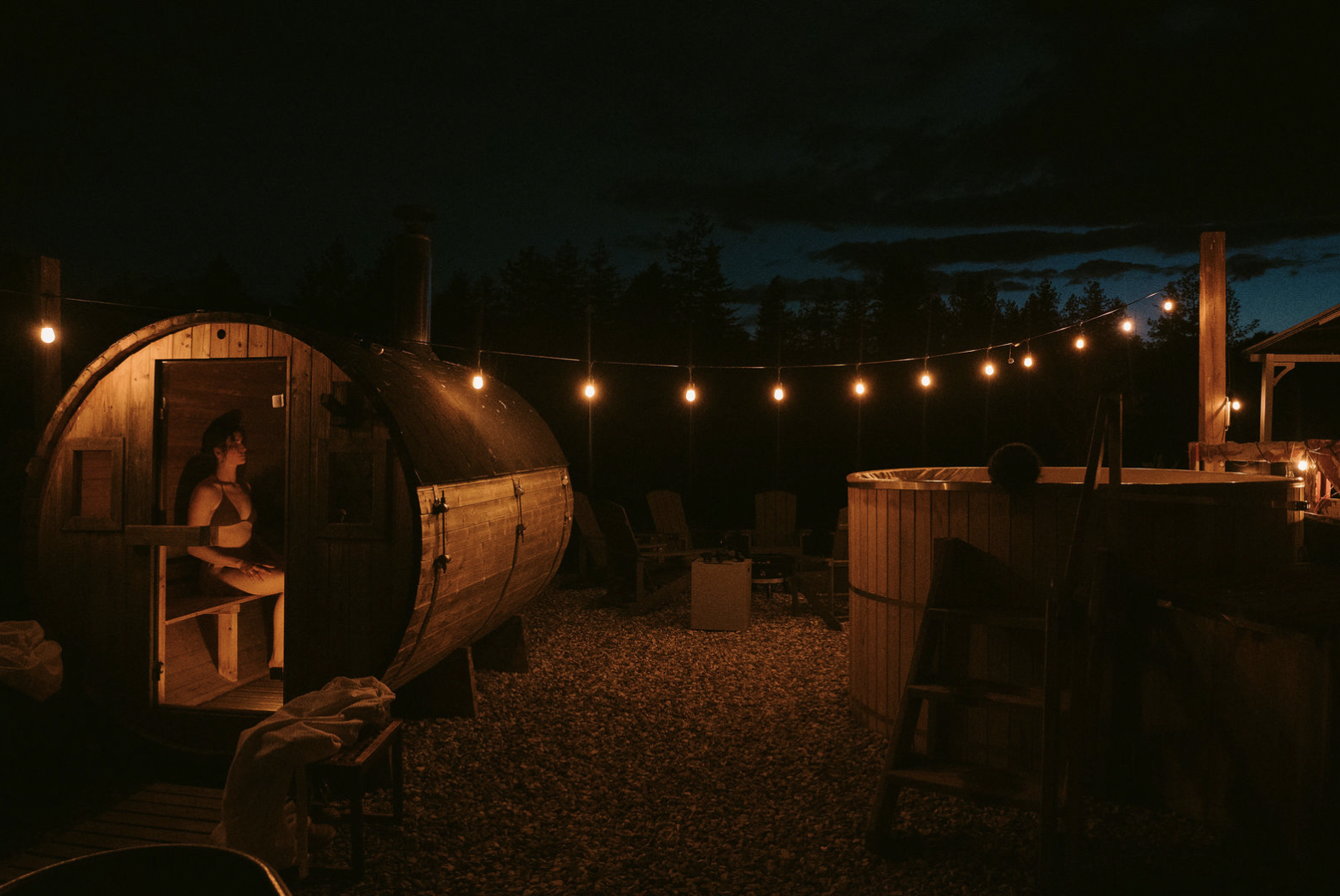 Nighttime scene with a woman inside a wooden barrel-shaped sauna, outdoor hot tub, string lights, chairs, and a firepit, dark sky with clouds in the background.