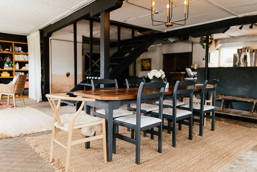 A dining area with a long wooden table surrounded by black chairs, a beige armchair, and a chandelier hanging from a white ceiling. There are shelves, a staircase, and other furnishings in the background.