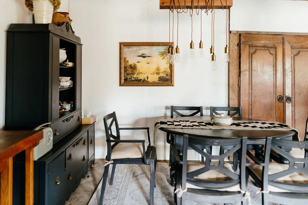 A dining room with a black sideboard, a wooden table with a black and white tablecloth, six black chairs with beige cushions, a framed painting of a UFO and a landscape, and a large wooden armoire.