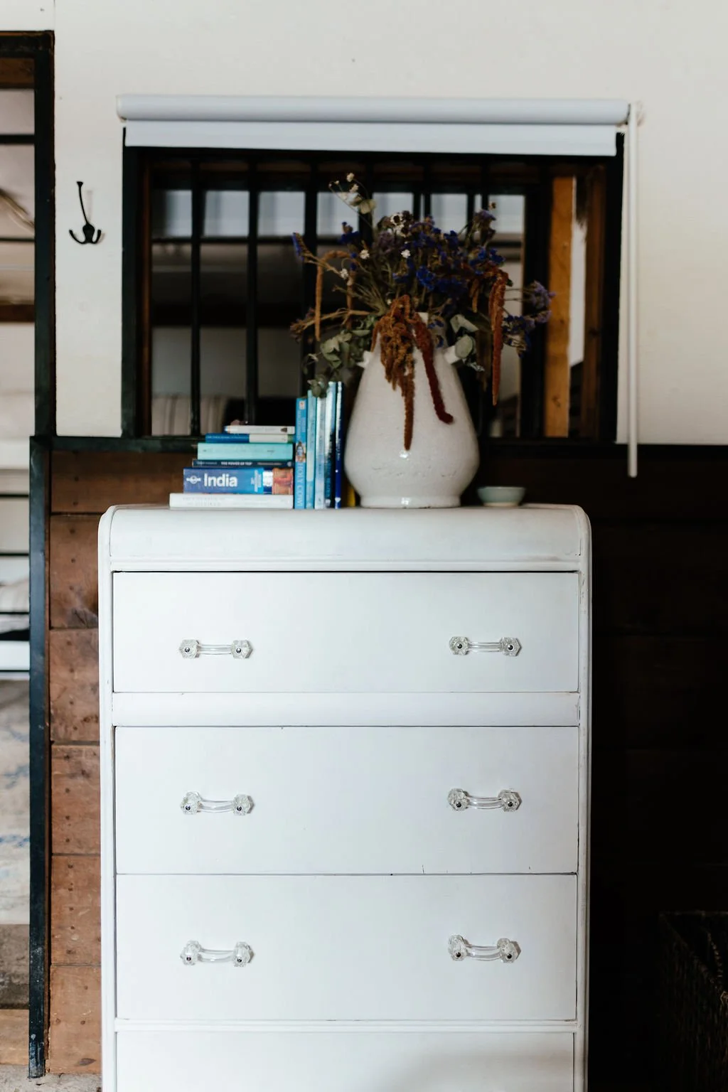 A white dresser with four drawers, topped with a large white vase holding dried flowers, and a small stack of blue and white books, situated in front of a window with black bars.