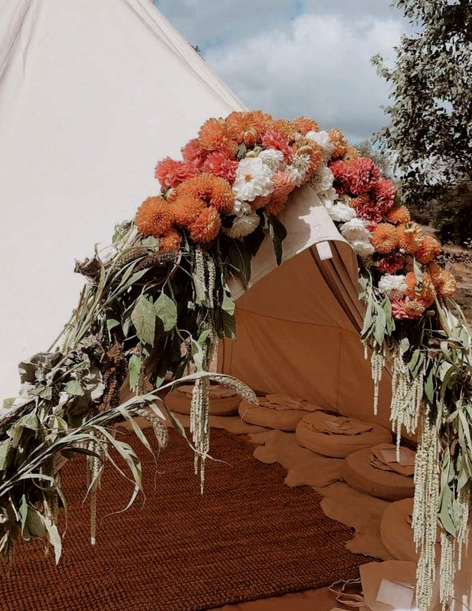 A wooden casket decorated with a large floral arrangement of orange, pink, and white flowers and greenery, placed outdoors near trees and a cloudy sky.