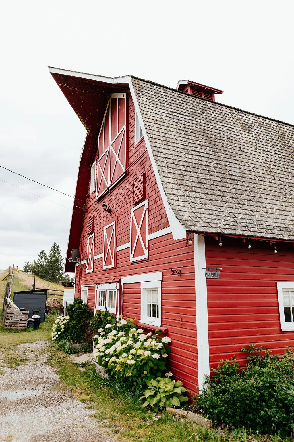 A red barn with white accents and a steep roof, surrounded by greenery and flowering bushes, under a cloudy sky.