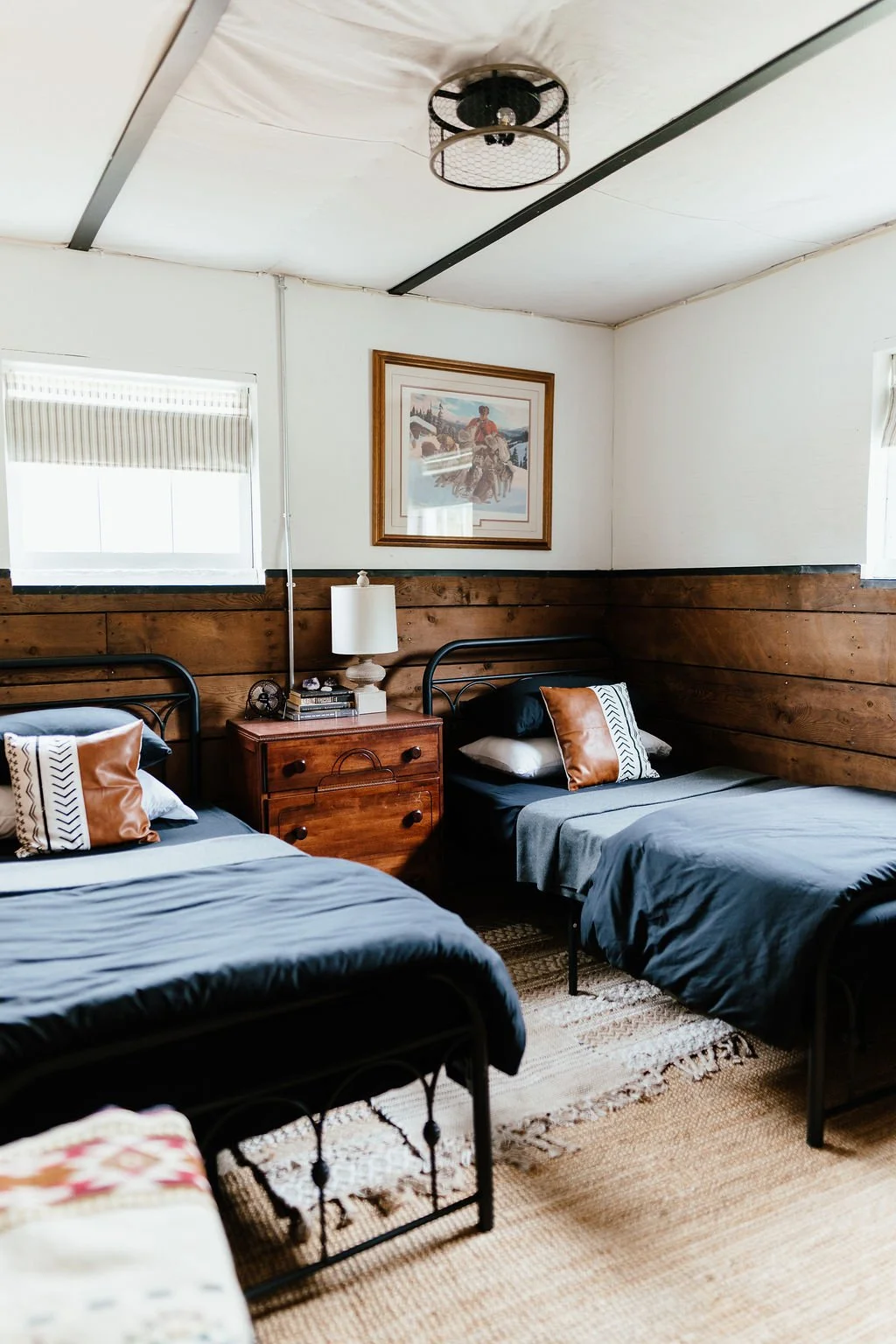 Room with two beds with black metal frames, grey bedding, and decorative pillows. A wooden nightstand between the beds holds a white lamp, books, and a small fan. The walls are half-white and half-wood paneling, with a framed picture of a landscape a