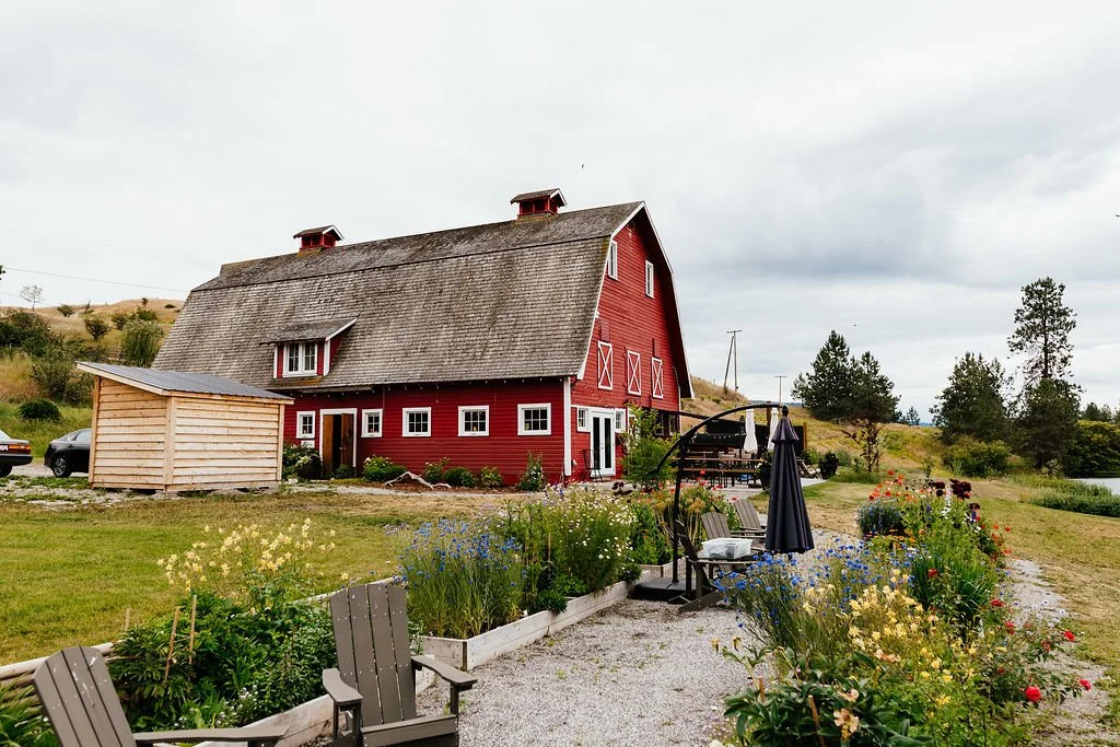 A red barn-style house with a steep roof, sitting in a garden area with colorful flowers, and patio furniture with a black umbrella.