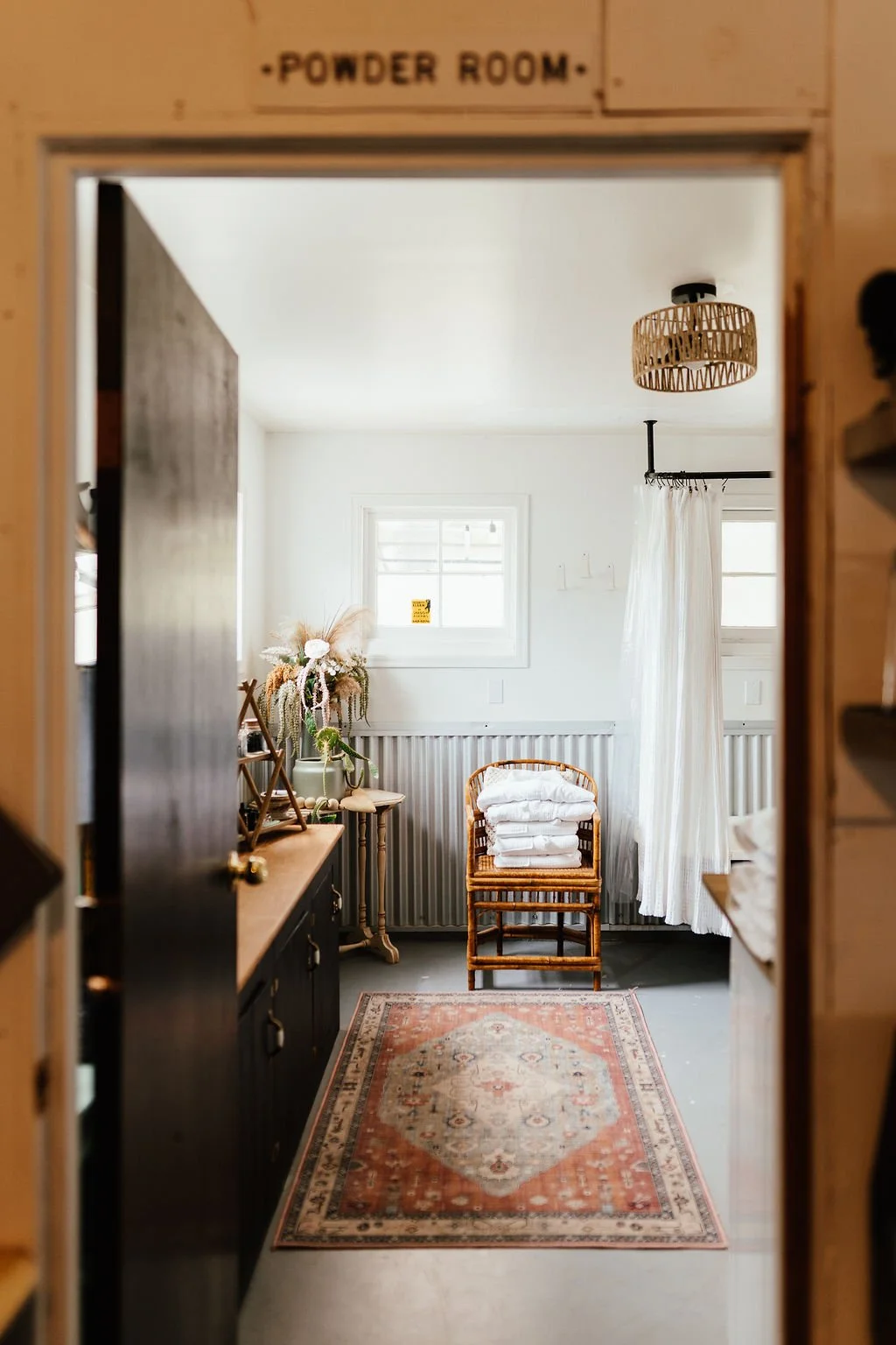 A cozy powder room with two small windows, white walls, and a white ceiling. The room features a wooden chair with white folded towels, a vintage-style beige and coral rug, and a black sideboard decorated with a floral arrangement. There is also a wh