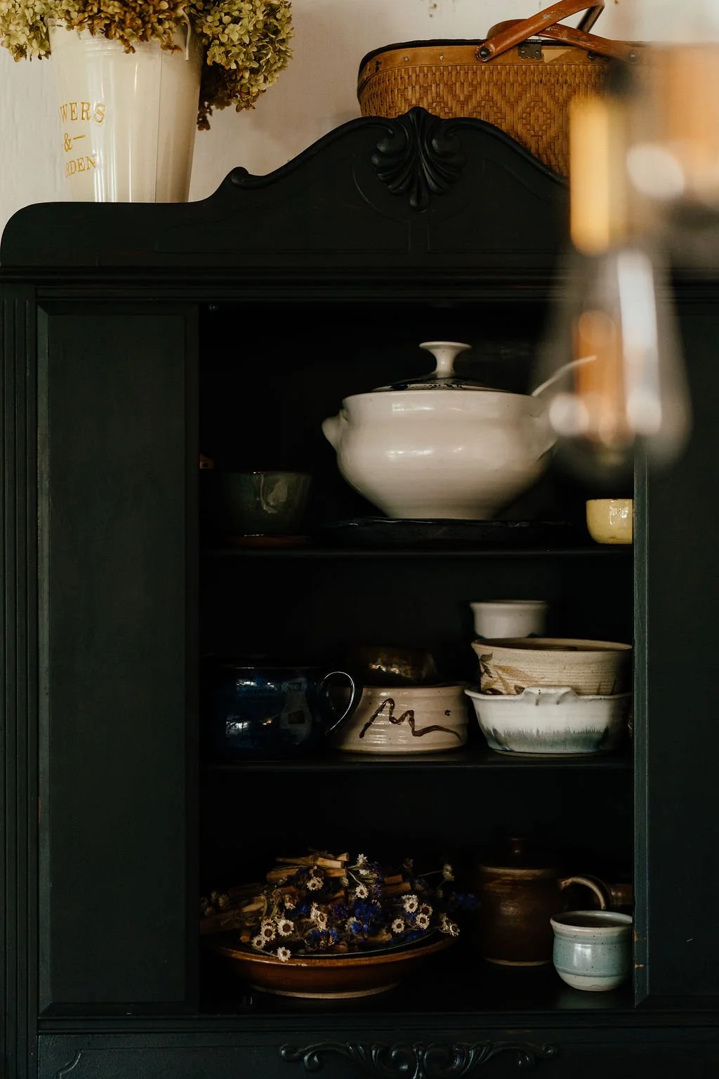 Interior of a black cabinet with shelves containing various ceramic and glass bowls and cups, a white teapot, a woven basket, a flower arrangement, and other decorative items.