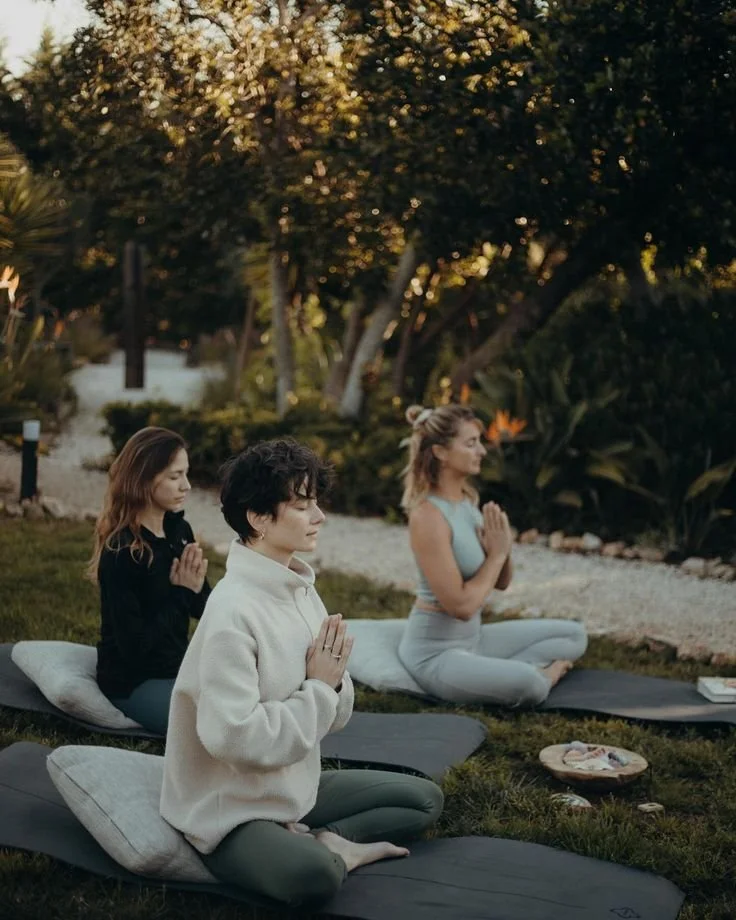 Three women practicing meditation outdoors on yoga mats in a lush garden at sunset.