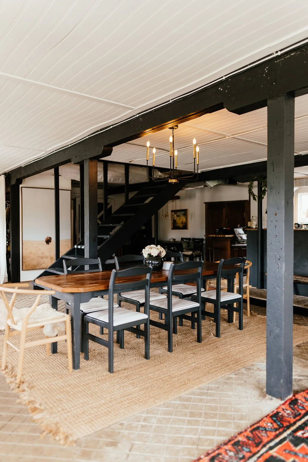 A dining room with a wooden table and six black chairs on a woven rug, a chandelier hanging from a white ceiling, and a staircase in the background.