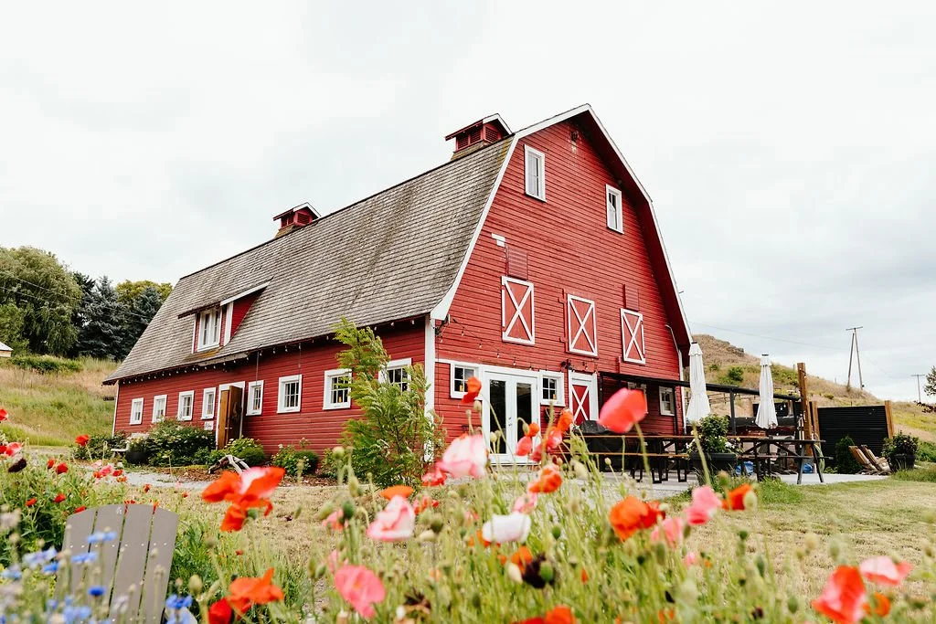 A large red barn with white trim and a sloped roof, surrounded by colorful flowers and outdoor seating, with cloudy sky and hilly landscape in the background.