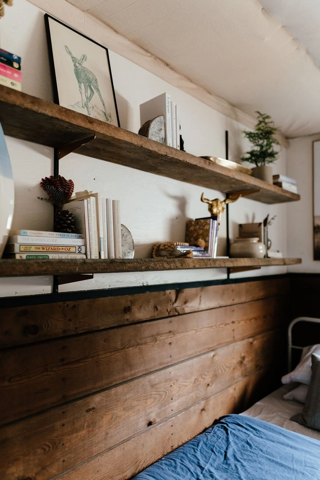 Wooden shelves decorated with books, framed artwork of a deer, potted plant, Buddha statue, and decorative objects in a cozy bedroom.