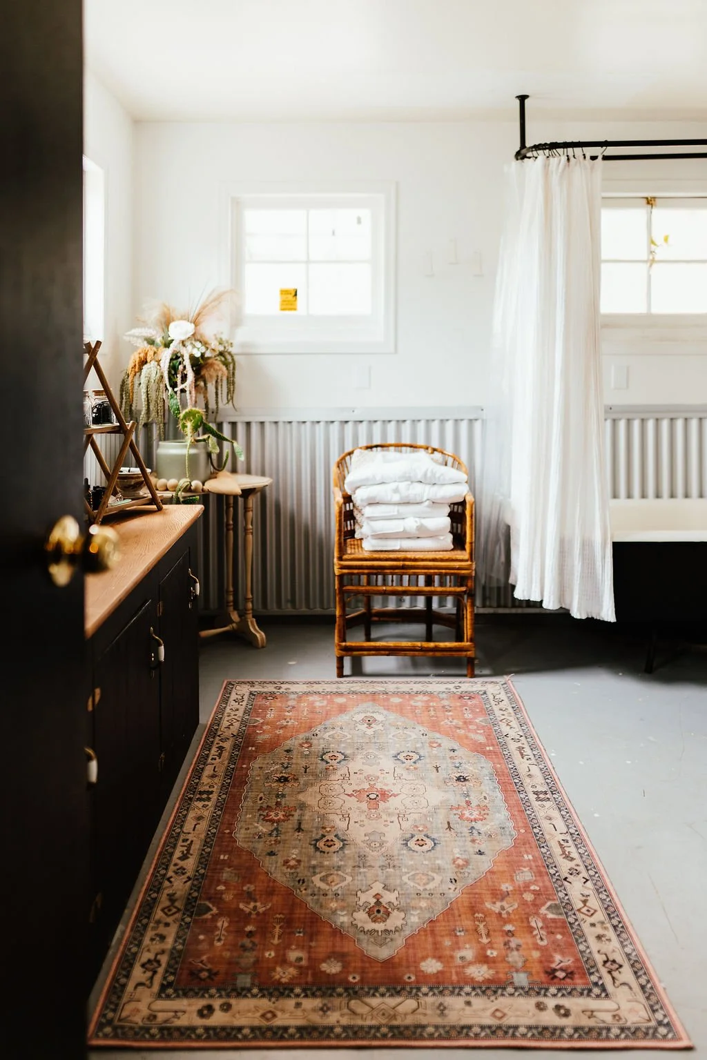 A cozy bedroom corner with natural light, a vintage rug, a bamboo chair with folded white linens, a wooden side table with decorative items, and a bed partially visible with a white curtain.