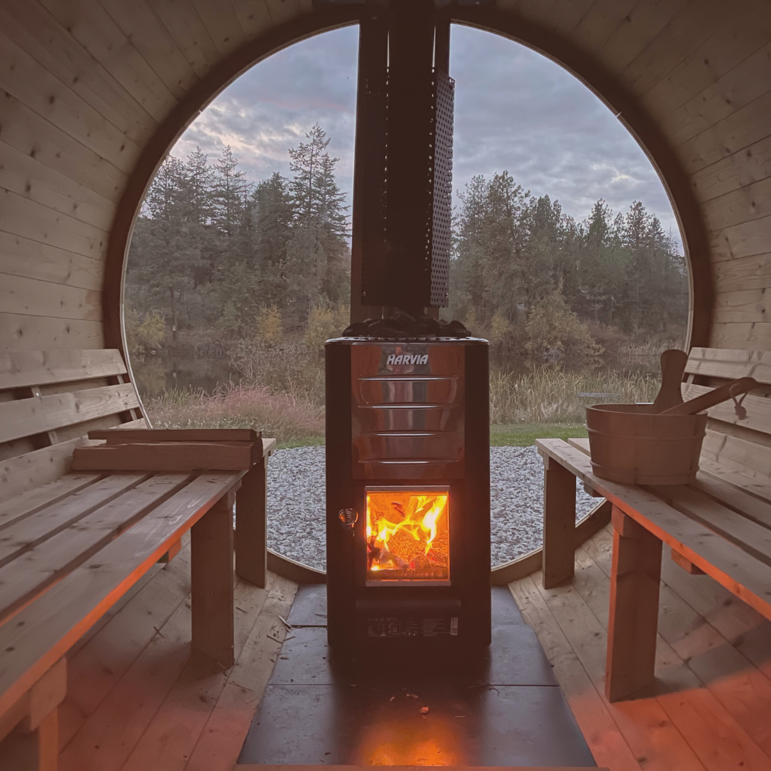 Inside a cozy wooden sauna with two benches, a fire stove in the center, and a large round window showing an outdoor landscape with trees and a cloudy sky.