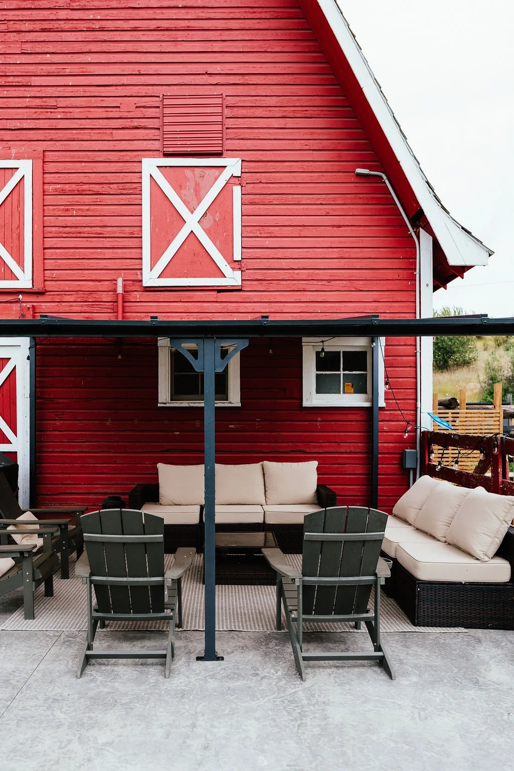 Outdoor patio area in front of a red wooden barn with white accents, including a seating area with couches and chairs, and a black metal railing overhead.