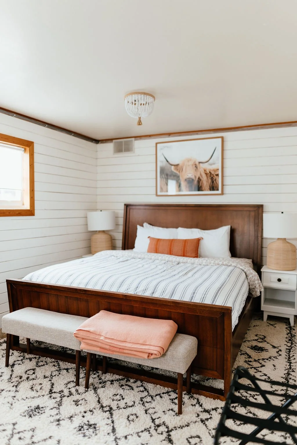 A bedroom with a wooden bed frame, white wood-paneled walls, and a framed picture of a highland cow with long horns. There are two white nightstands with beige lamps, a window with wooden trim, a patterned black and white rug, and a bench at the foot