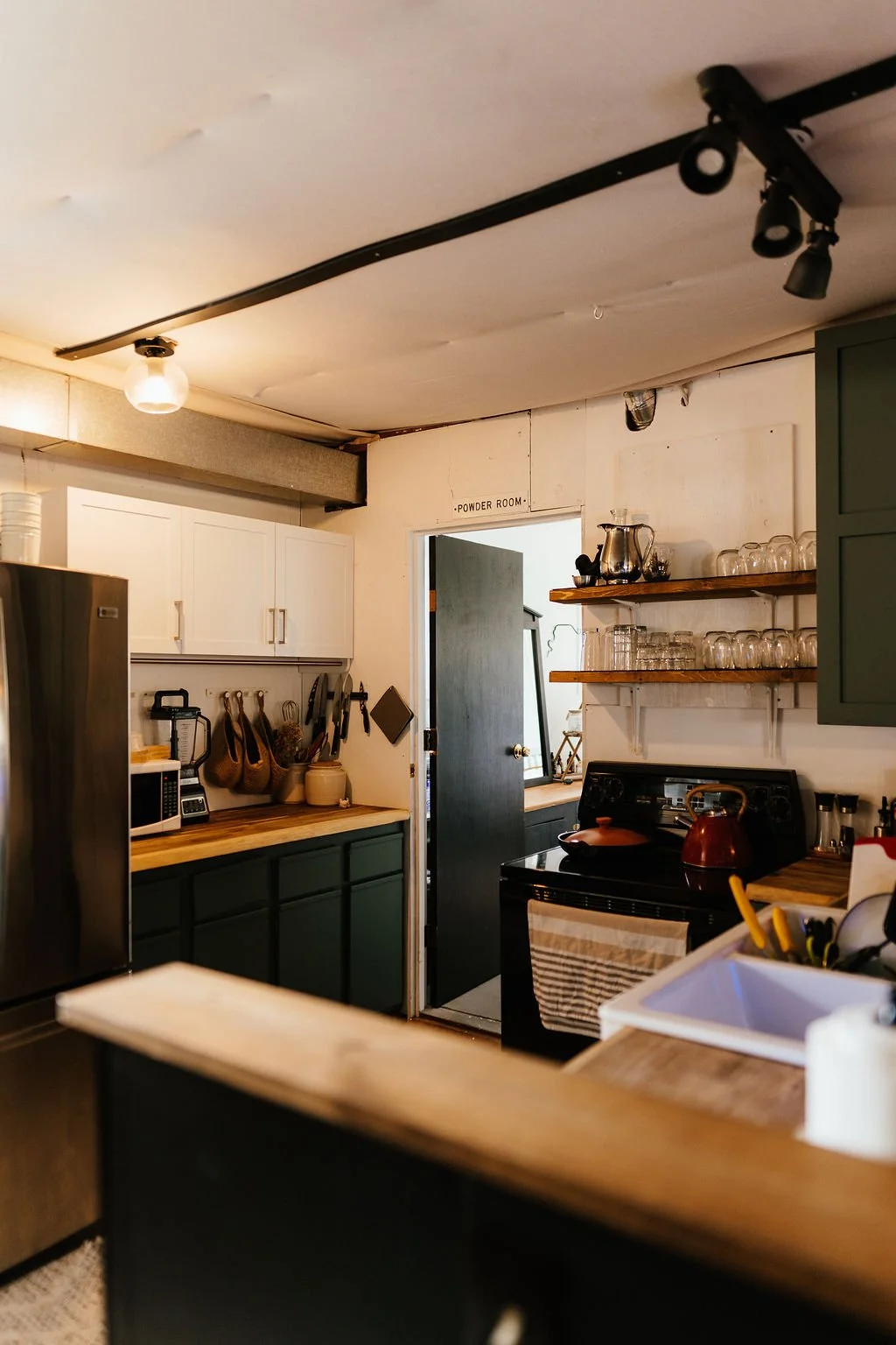 Cozy kitchen with white upper cabinets, black lower cabinets, wooden countertops, open shelves with glasses and a silver pitcher, black stove with a red kettle, and a doorway labeled 'Powder Room' leading to another room.
