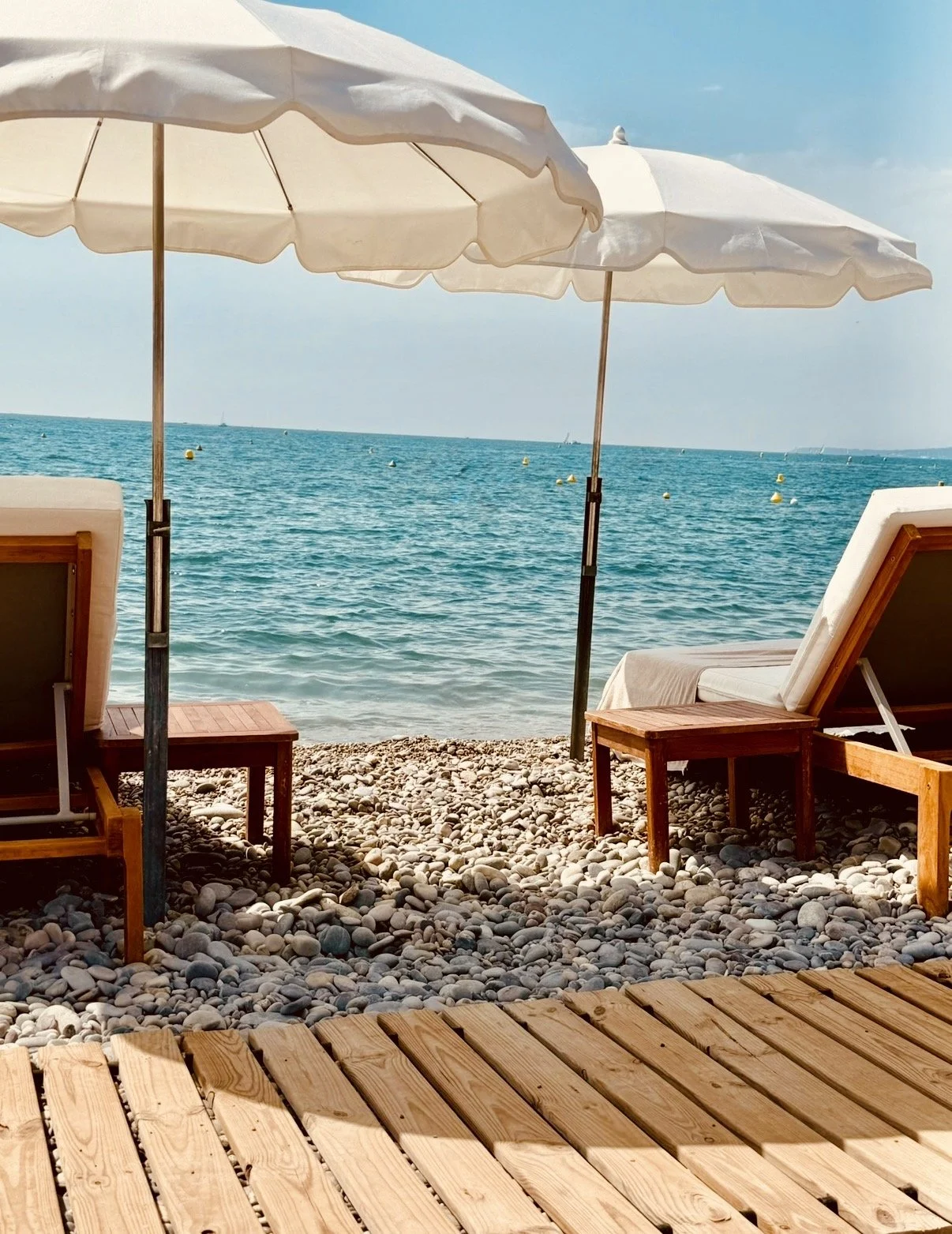 Wooden lounge chairs and white umbrellas overlooking the Mediterranean Sea at a beach club.