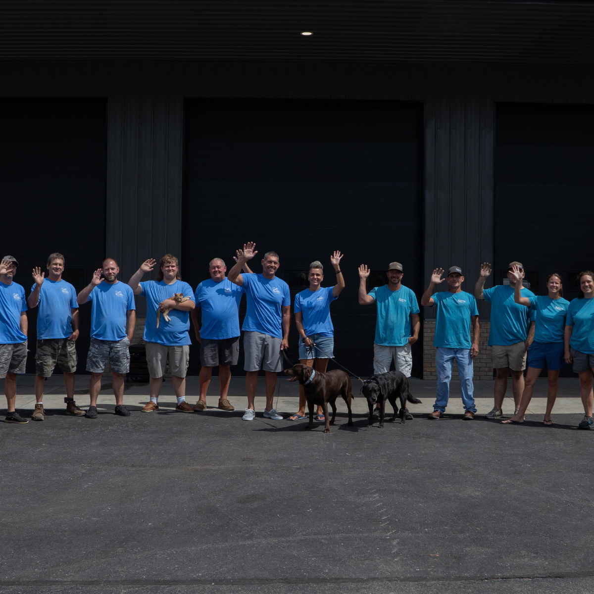 Group of people wearing matching blue shirts, standing outdoors in front of a black building, waving at the camera, with two dogs on leashes.