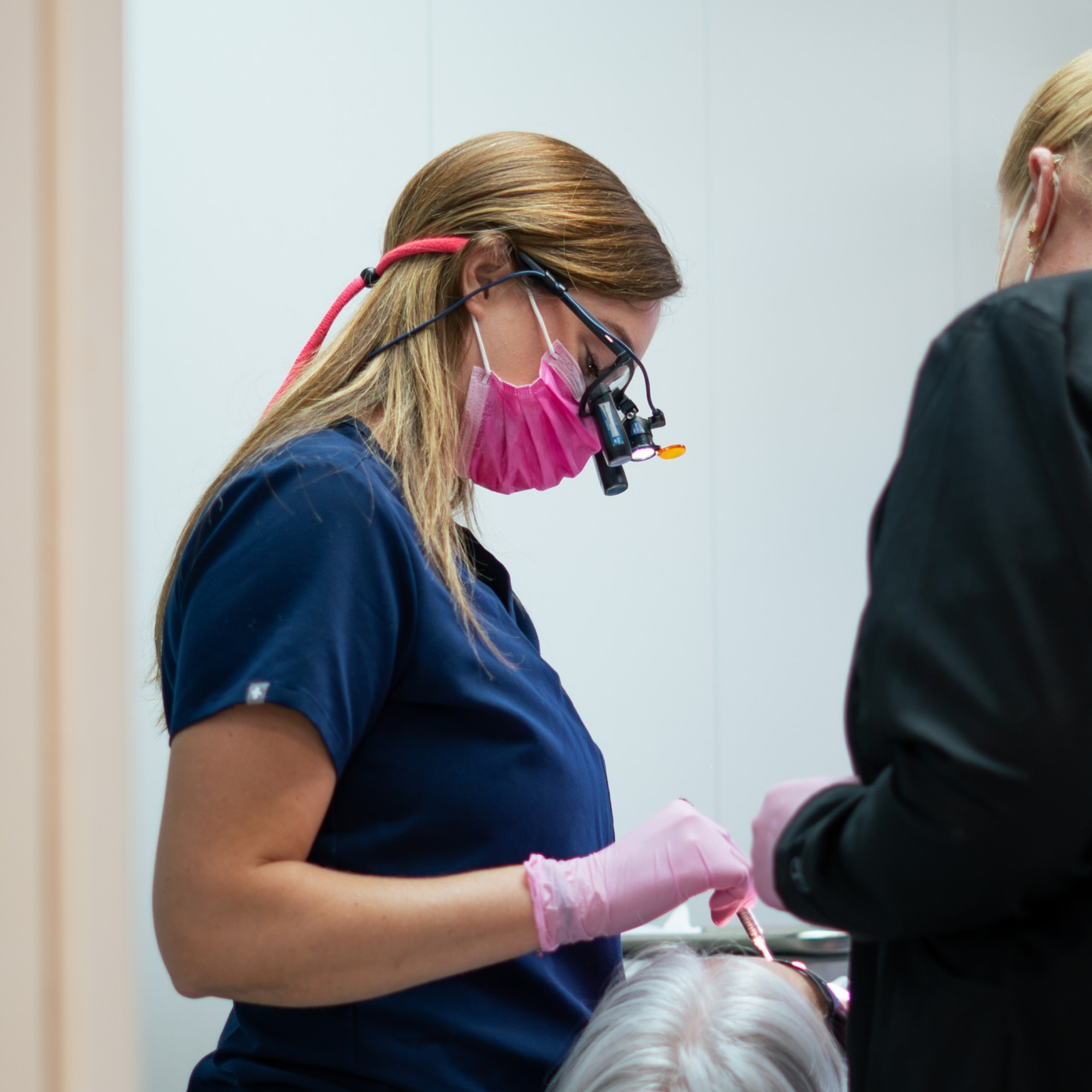 Dental professional wearing safety glasses and a pink mask working on a patient's teeth with dental tools in a clinical setting.