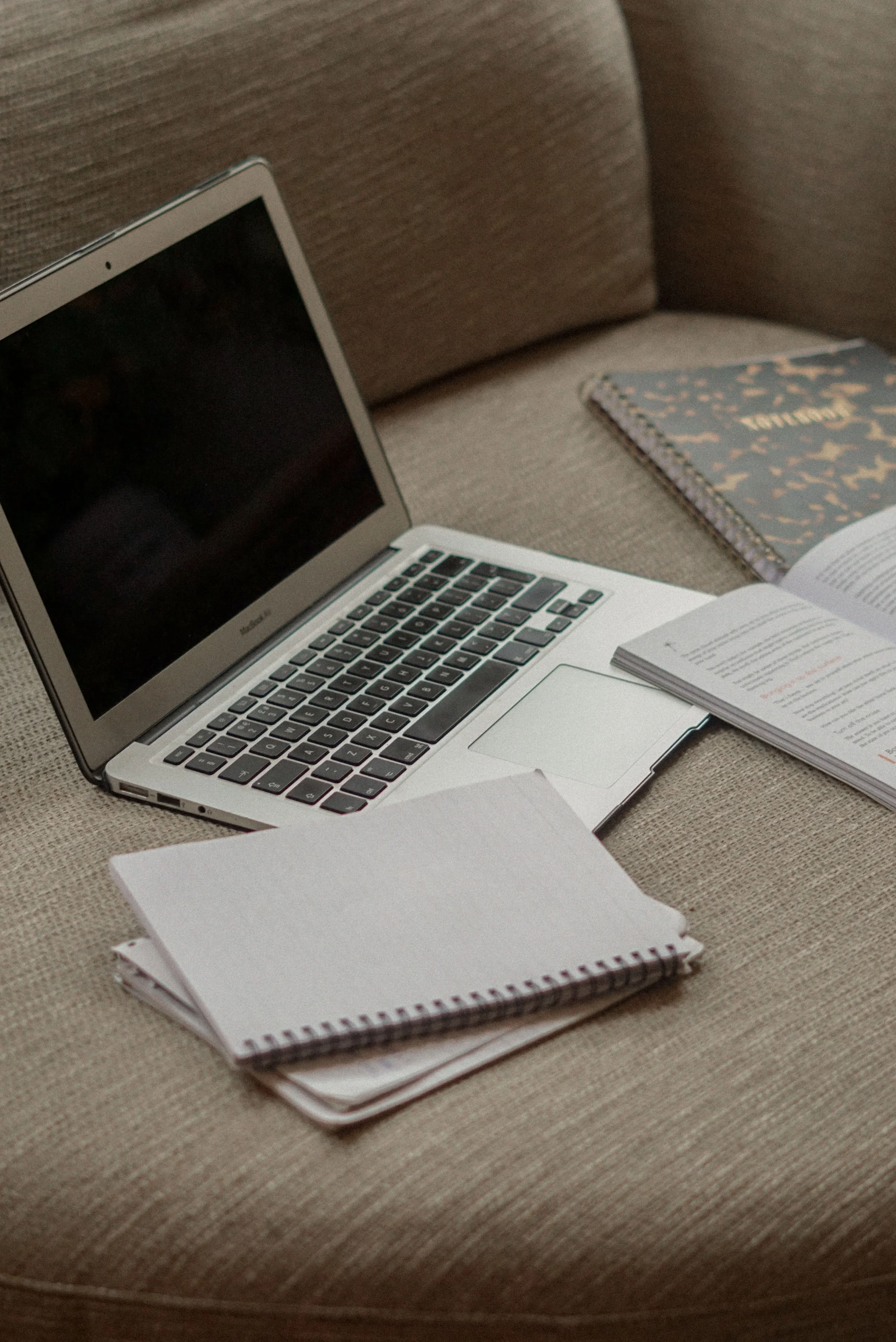 A MacBook laptop, open and turned on, sitting on a textured beige surface with a spiral notebook and an open book nearby.