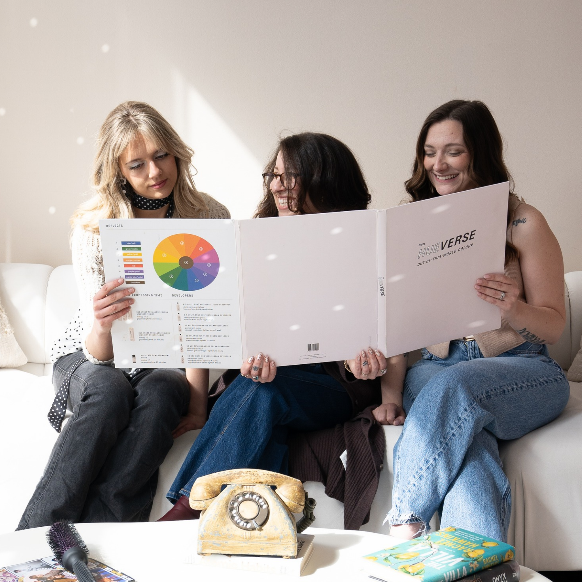 Three women sitting at a table, looking at a large book with a color wheel on the page. An antique rotary telephone is on the table, along with a book titled 'The Yellow House' and some magazines. A vintage microphone and a hairbrush are also visible.