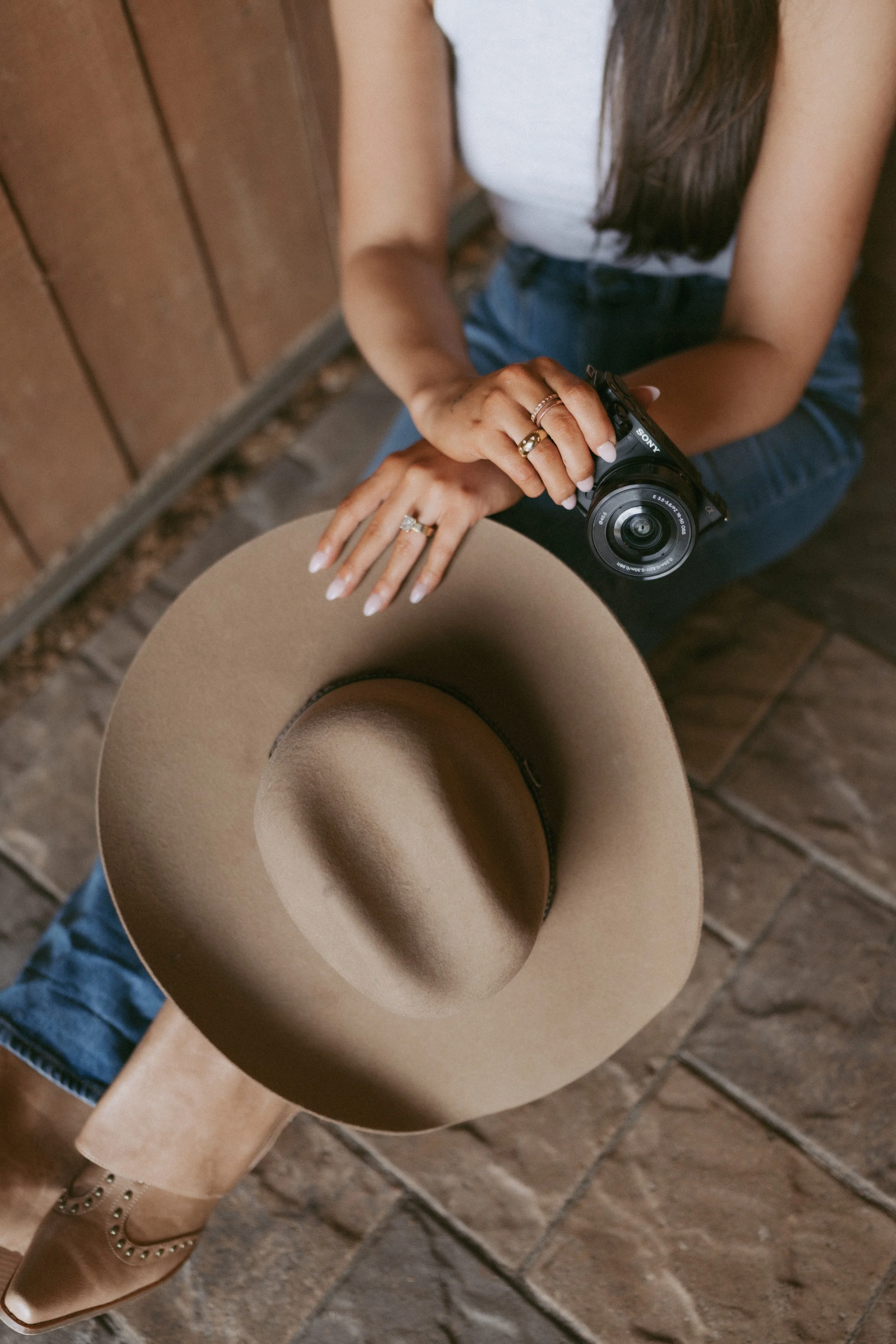 A woman sitting on a wooden bench, holding a camera, with a wide-brimmed beige hat on her lap, wearing rings and a sleeveless top, and wearing brown ankle boots with laces and small heel.