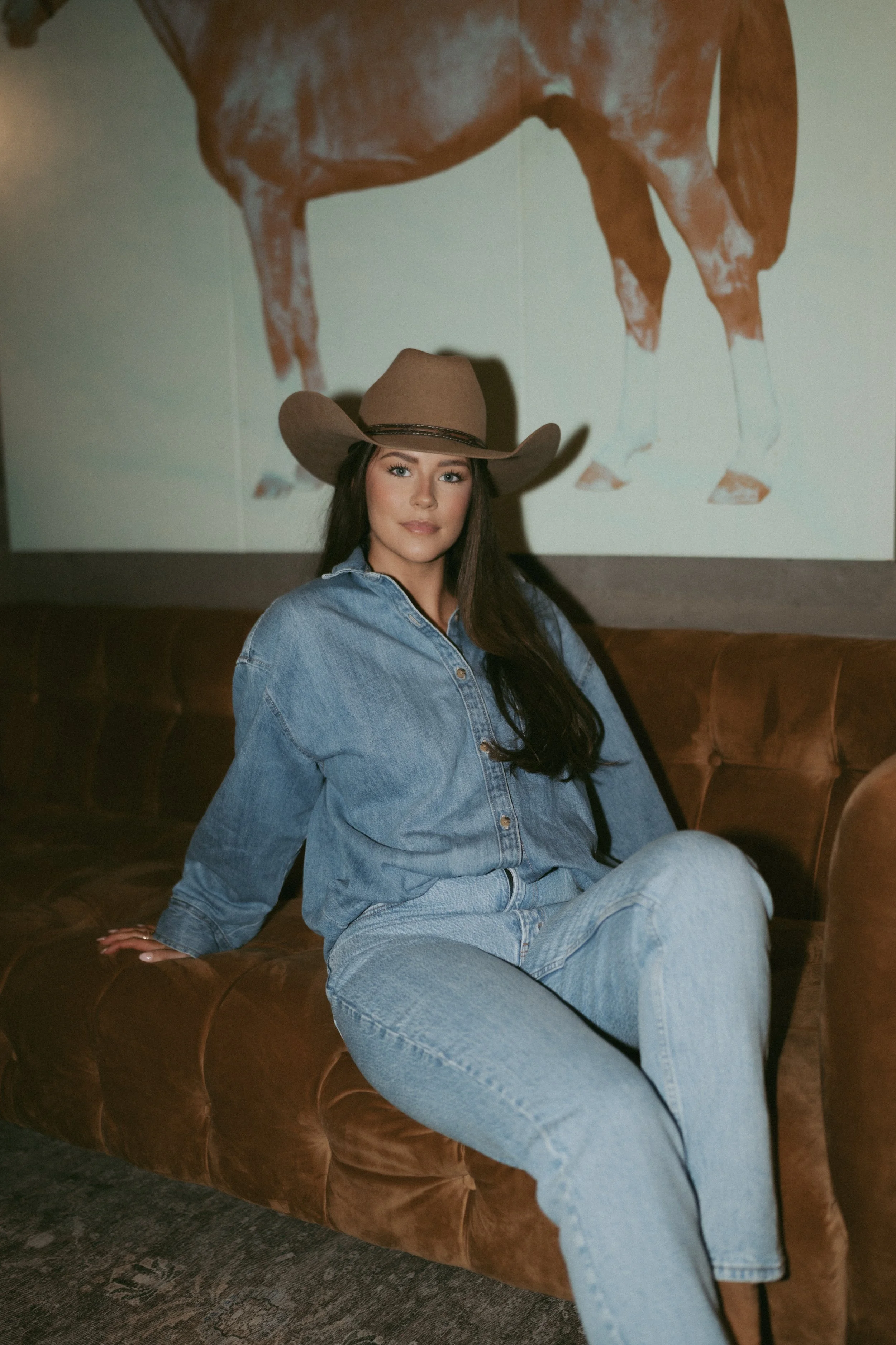 A young woman with long dark hair and fair skin wearing a tan cowboy hat, denim shirt, and matching jeans is sitting on a brown leather sofa. She is in a relaxed pose with one arm resting on the sofa. There is a large wall art of a close-up of a brown horse's body and legs behind her. The setting appears to be indoors with a casual, rustic vibe.