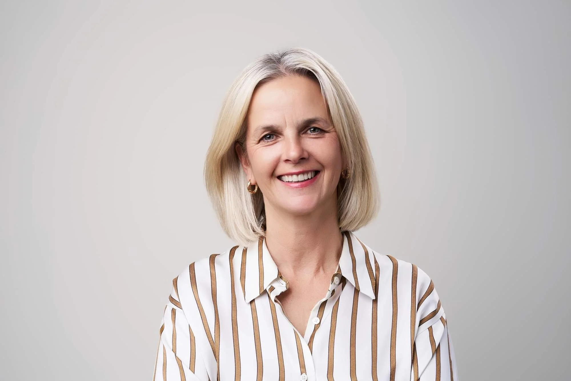 A smiling female interior stylist with shoulder-length blonde hair, wearing a white blouse with brown vertical stripes, standing against a plain light gray background.