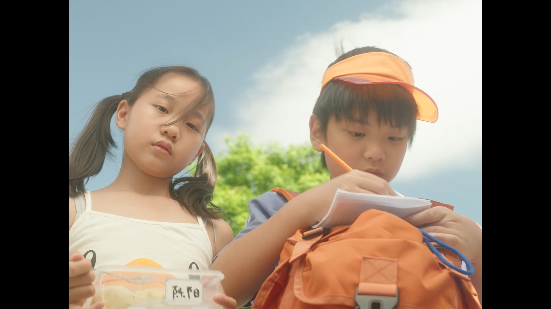 A young girl with pigtails and a young boy wearing an orange visor and backpack, both outdoors on a sunny day, with the boy writing in a notebook and the girl holding a container of food.