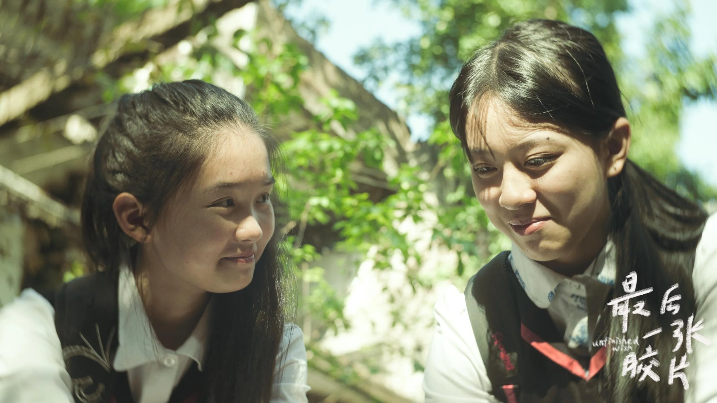 Two young girls with long dark hair and light skin looking at each other outdoors in a green, leafy environment.