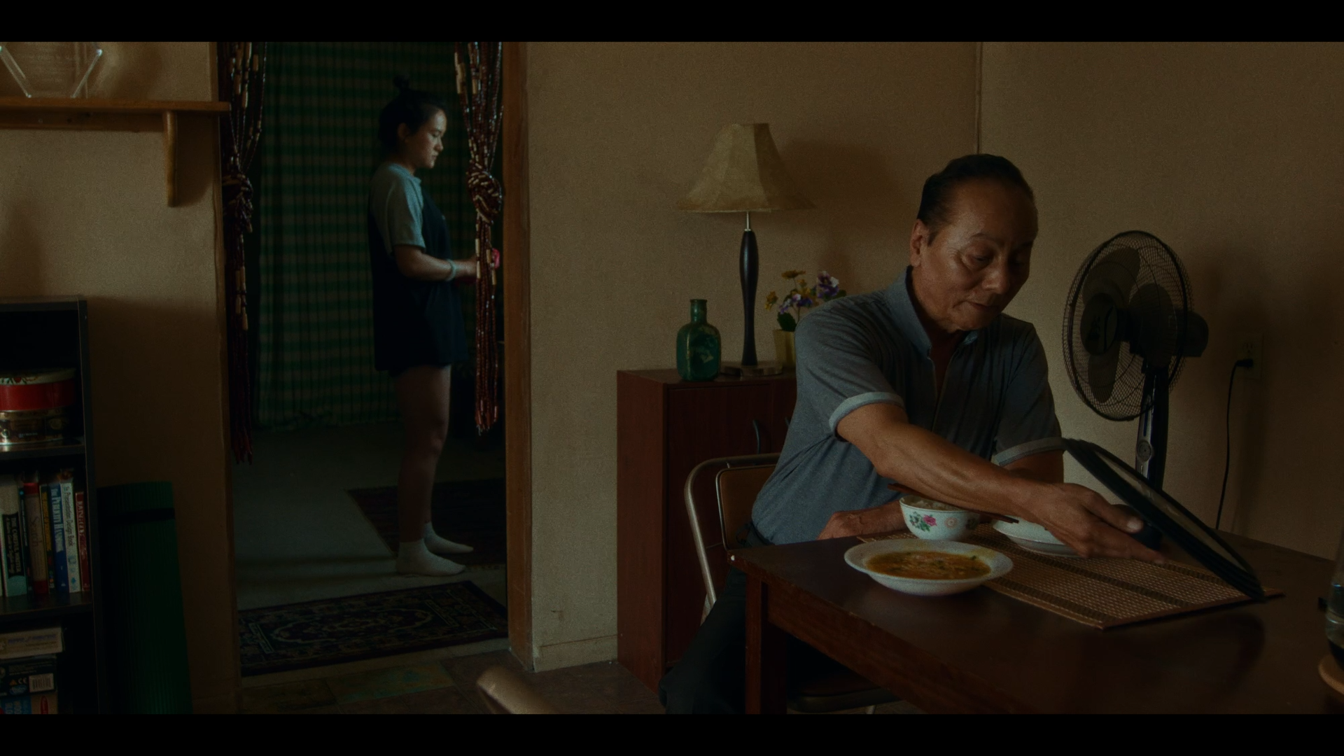 A woman stands in a doorway looking at her phone, while an older man sits at a table reading a book, with a bowl of soup and a bowl on the table, in a dimly lit room with a fan and decorative items.