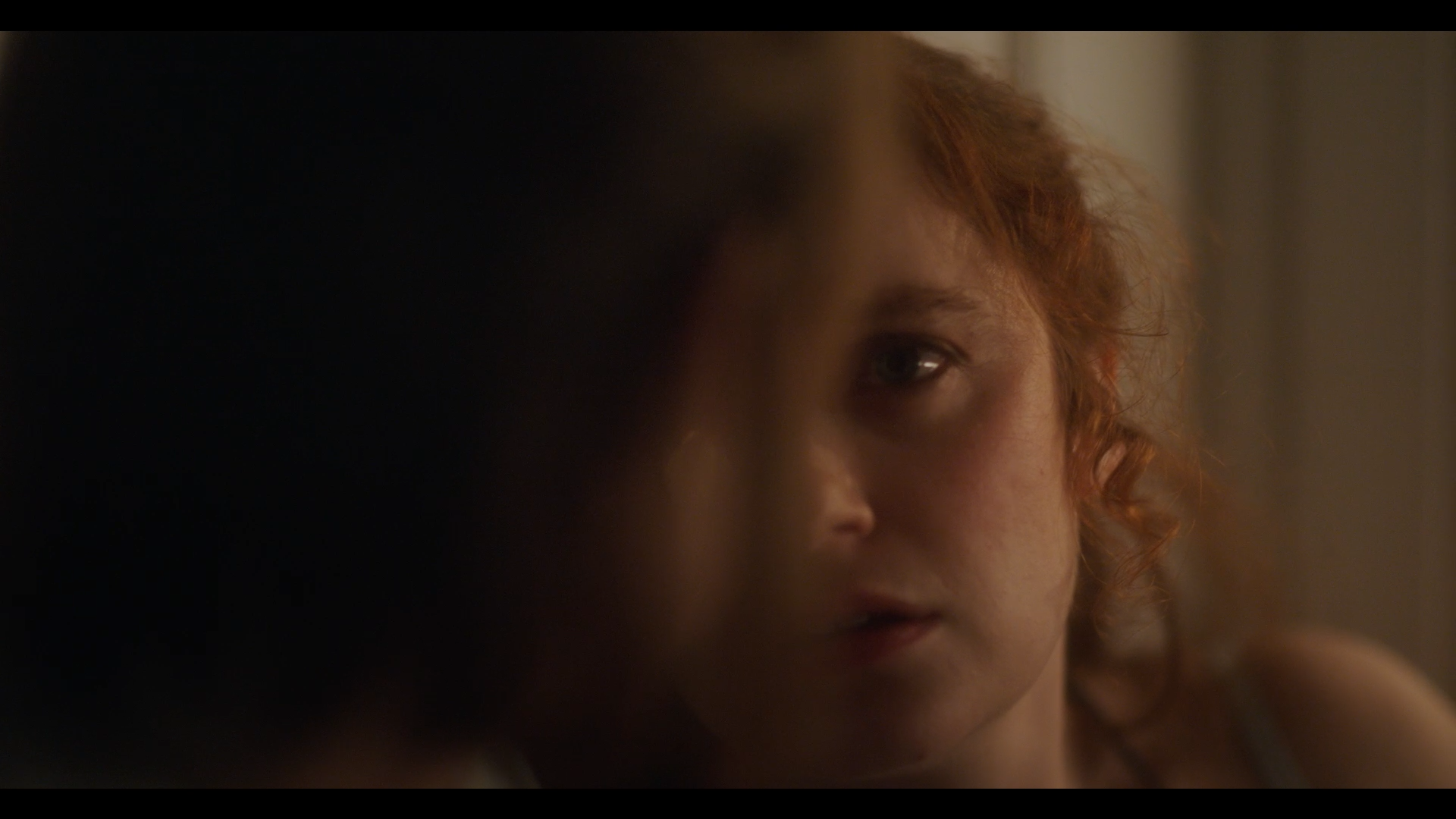 Close-up of a woman with red curly hair and intense expression, partially obscured by a shadow.
