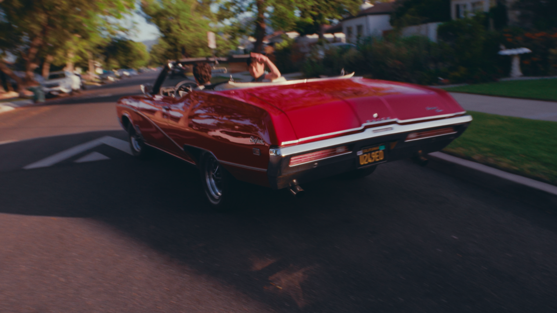 A red vintage convertible car driving down a suburban street with two people inside, one waving and another looking back, surrounded by trees and houses.