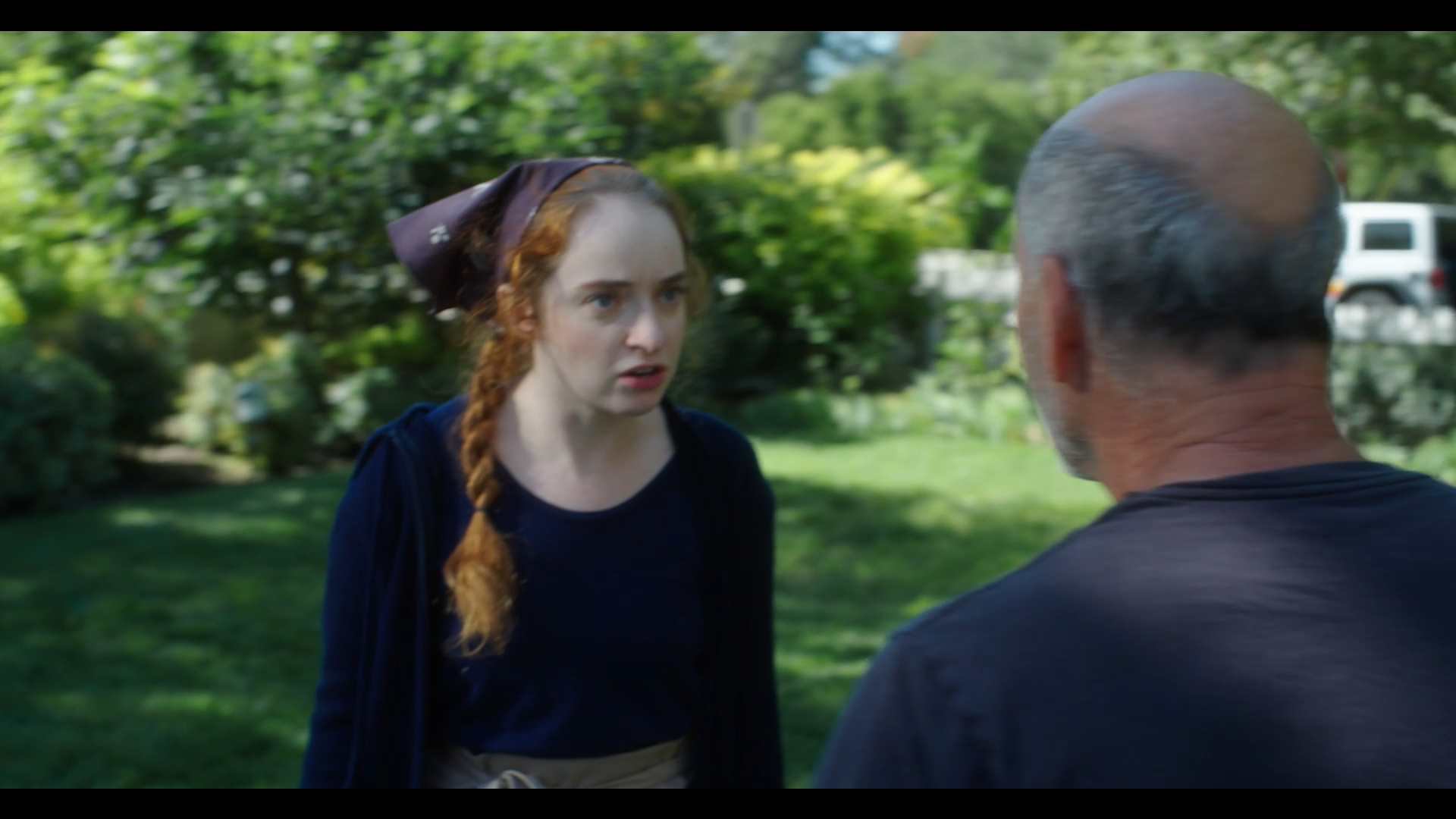 A woman with red hair in a braid and wearing a dark blue outfit is talking to a man with gray hair and glasses in a grassy outdoor setting with trees and a white vehicle in the background.