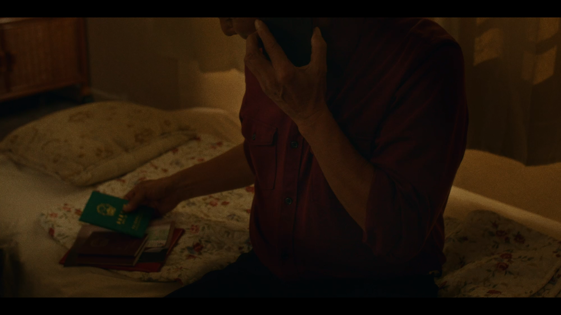 A person kneeling on a bed holding a green passport in one hand and a small stack of passports or documents on the bed, with a floral bedsheet and a pillow in the background.