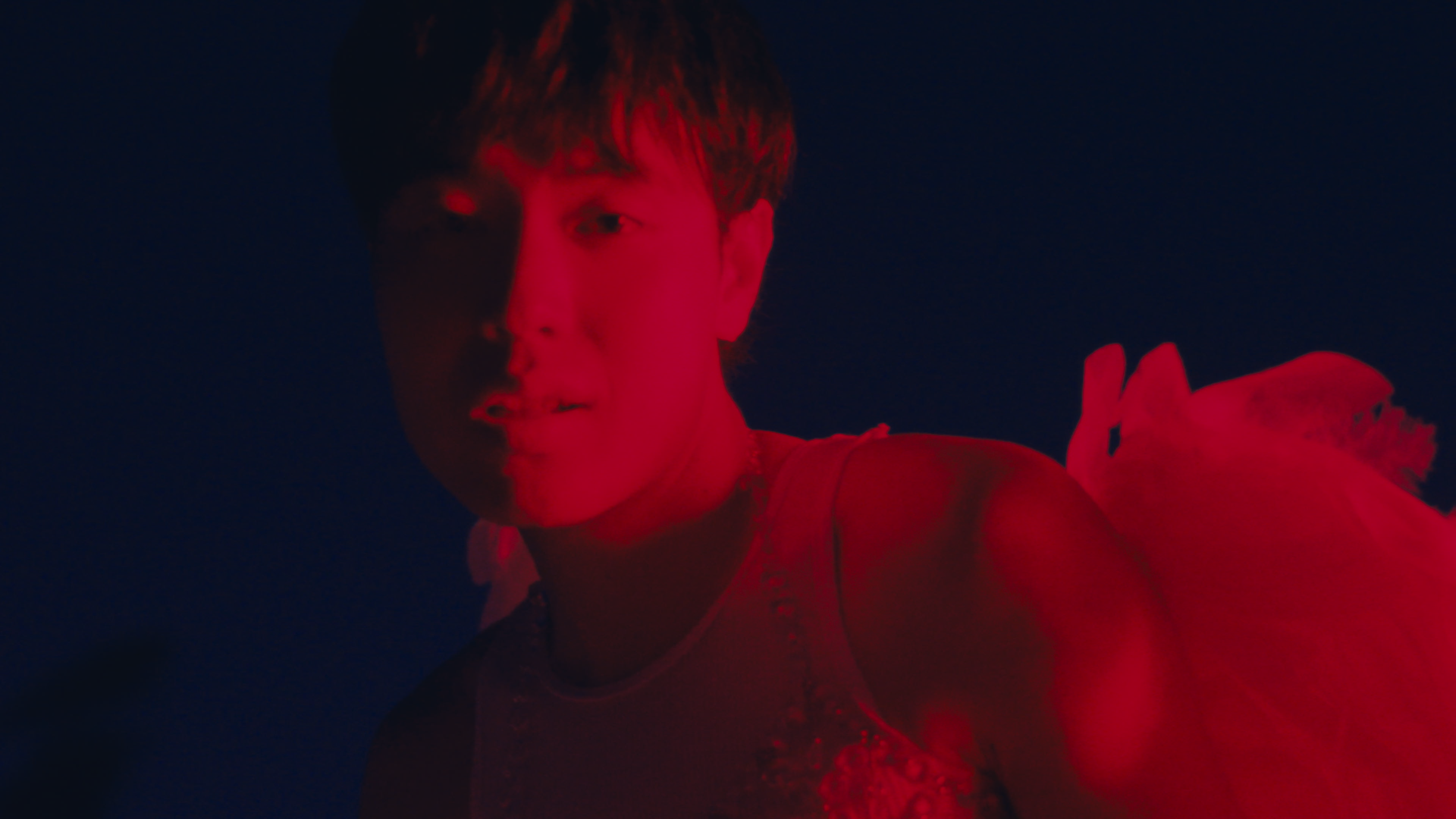 A young man with dark hair illuminated by red lighting, looking into the camera with a slight smile, against a dark background.
