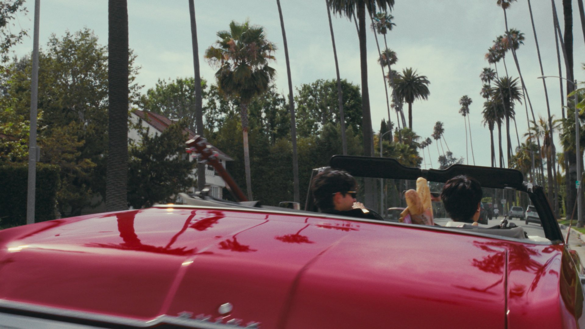 People riding in a red convertible car on a street lined with tall palm trees, with a cloudy sky overhead.