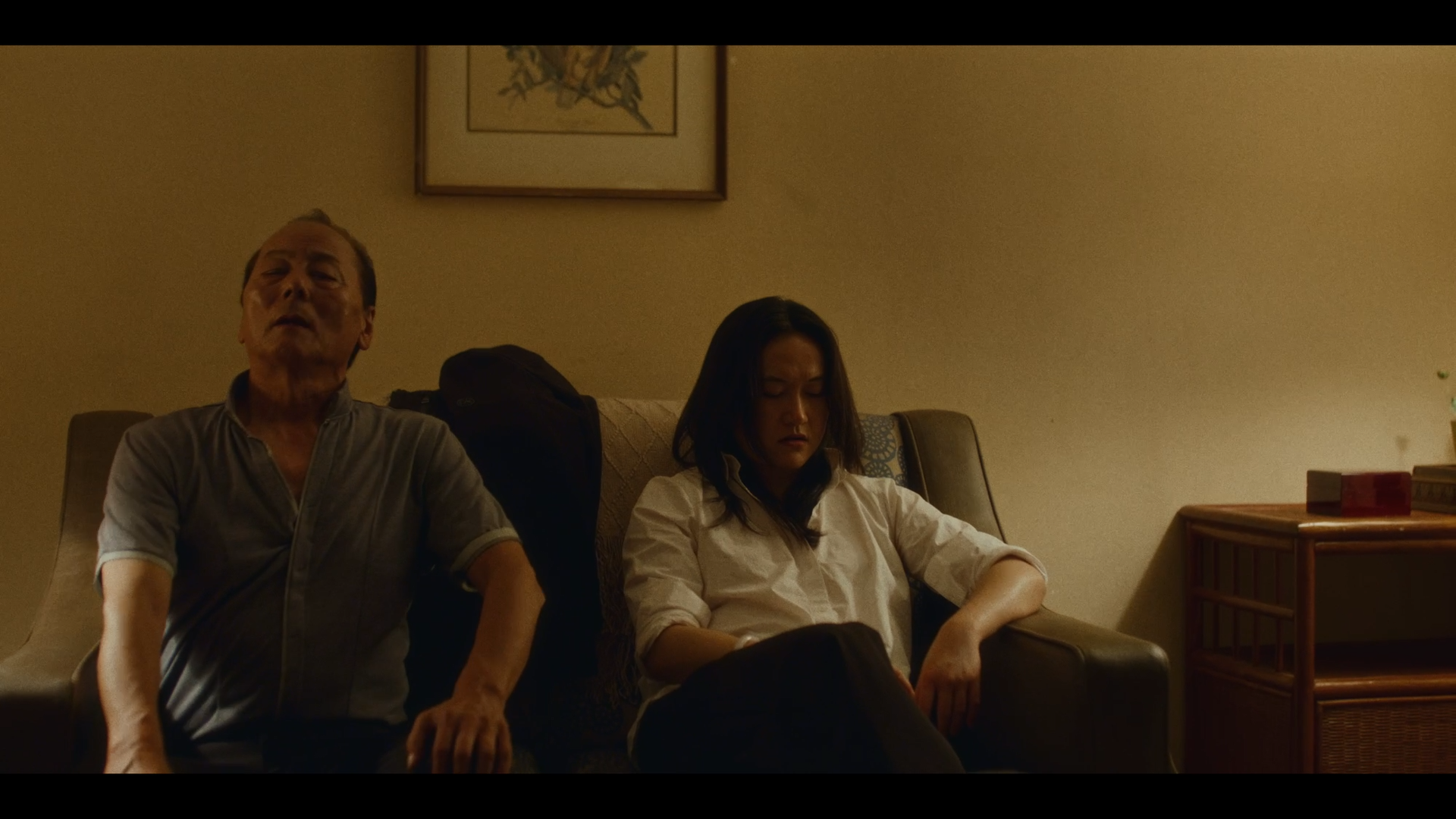 A man and a woman sitting on a sofa in a dimly lit room, with the man on the left and the woman on the right, appearing tired or contemplative.