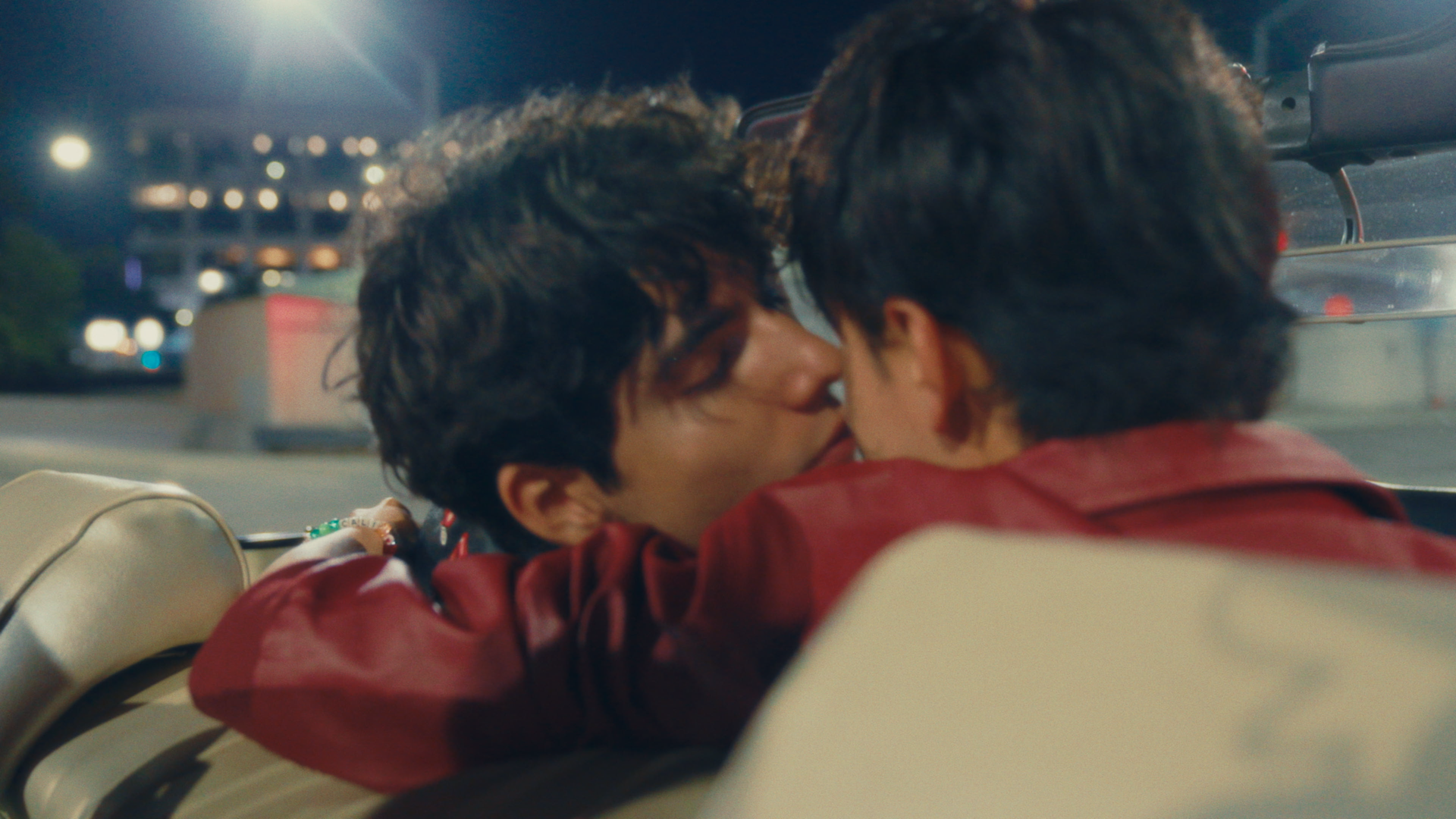 Two men with dark curly hair sharing a kiss in the backseat of a car at night, with city lights visible outside the window.