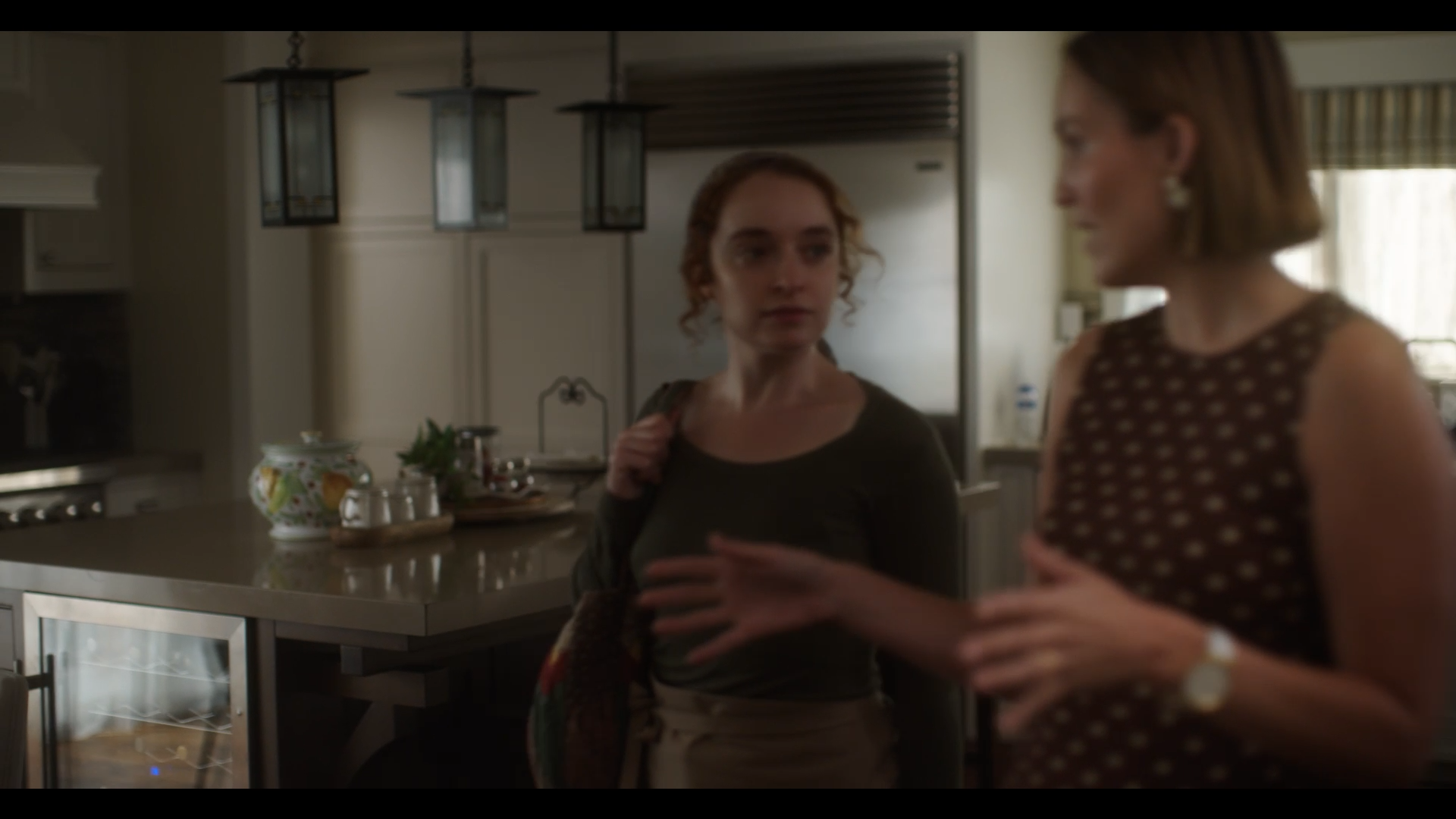 Two women are talking in a kitchen. The woman on the left has curly red hair and is wearing a dark top, while the woman on the right has straight brown hair and is wearing a sleeveless, polka-dot top. There are hanging lamps and kitchen appliances in