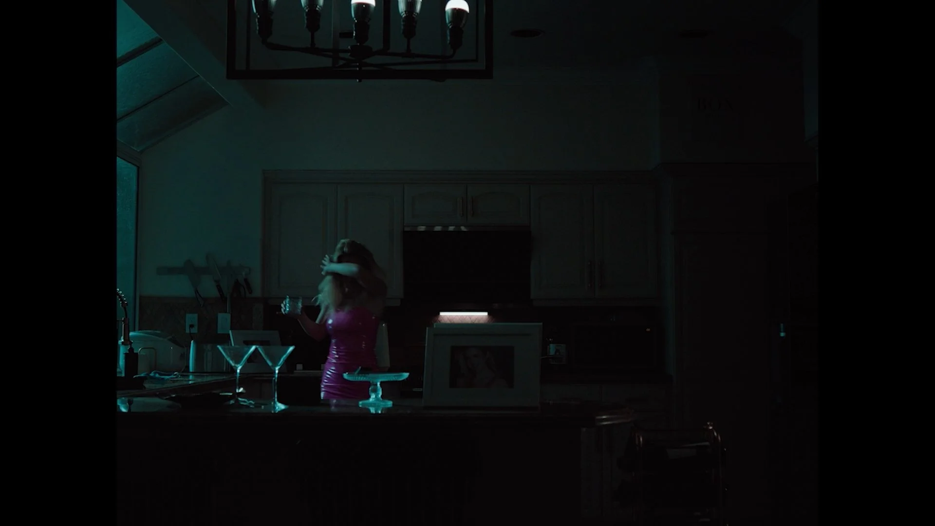 A woman in a pink dress standing in a dark kitchen, holding a drink and brushing her hair back. There are two martini glasses on the counter, and the room has dim lighting with a chandelier hanging from the ceiling.