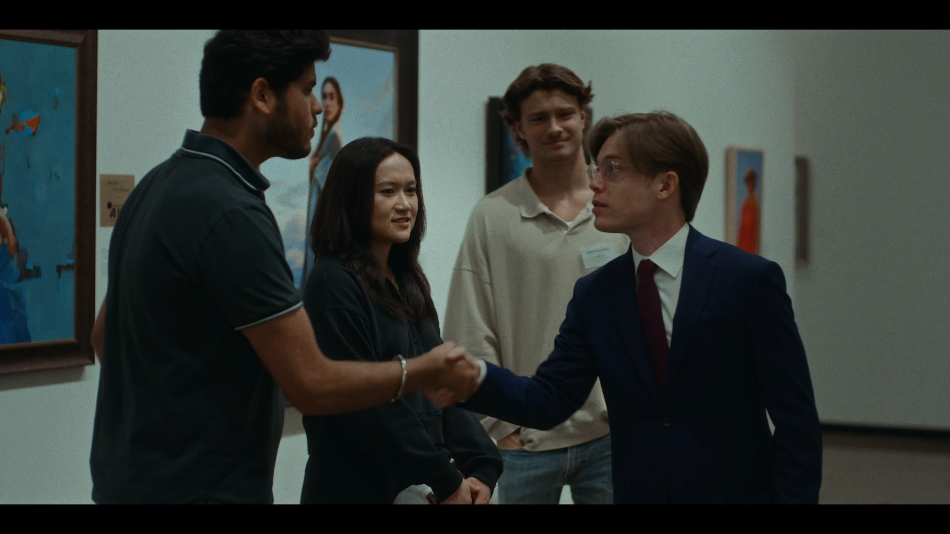 Two men shake hands in a room with art on the walls, while three other people stand nearby watching.