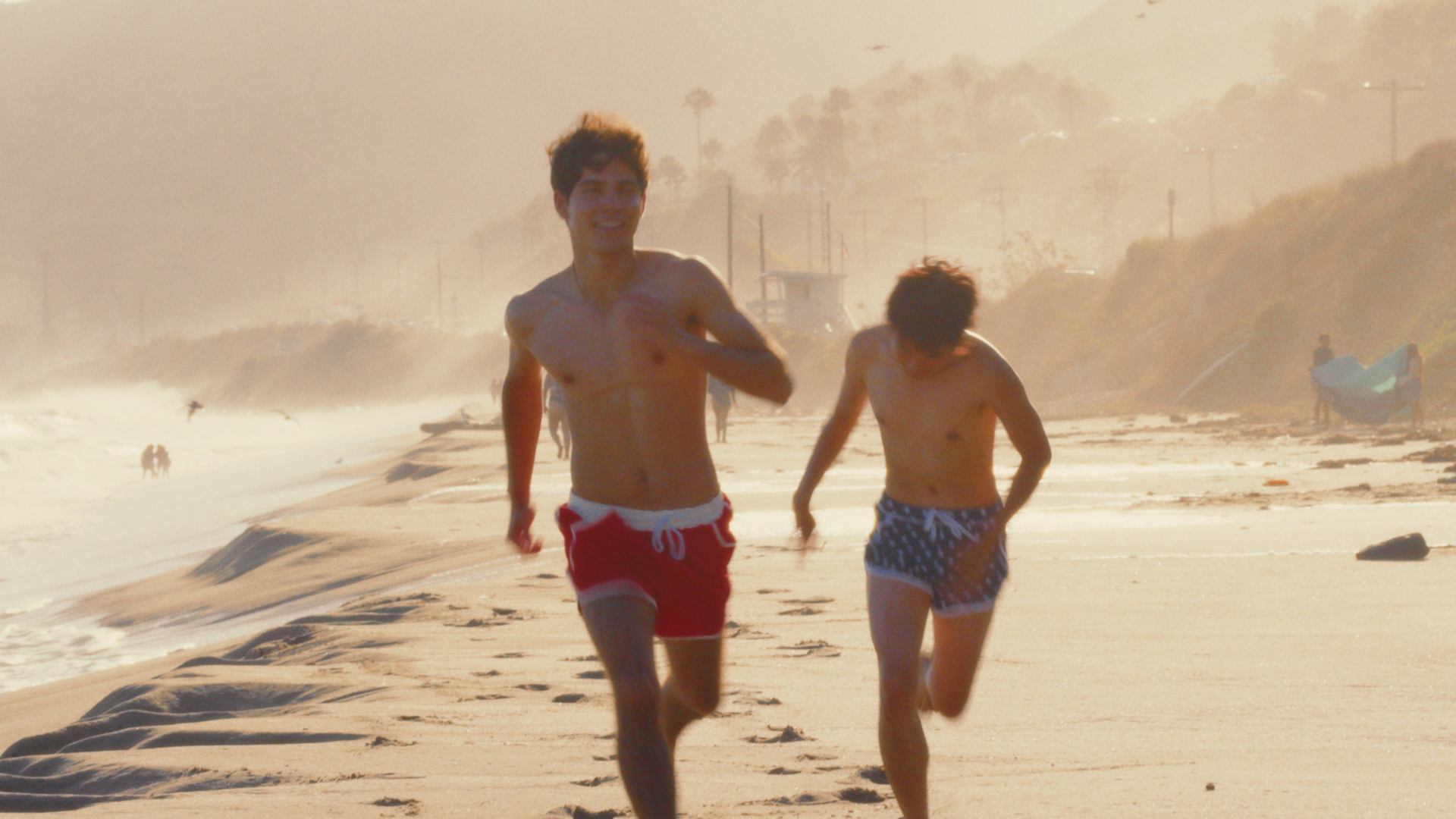 Two boys running along the beach in swimsuits, with waves crashing nearby and a sandy shoreline in the background.