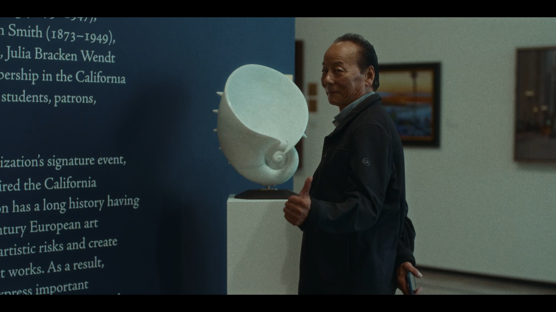A man stands in front of a blue informational display at a museum, giving a thumbs-up gesture, holding a small object in his left hand, with framed pictures hung on the wall in the background.