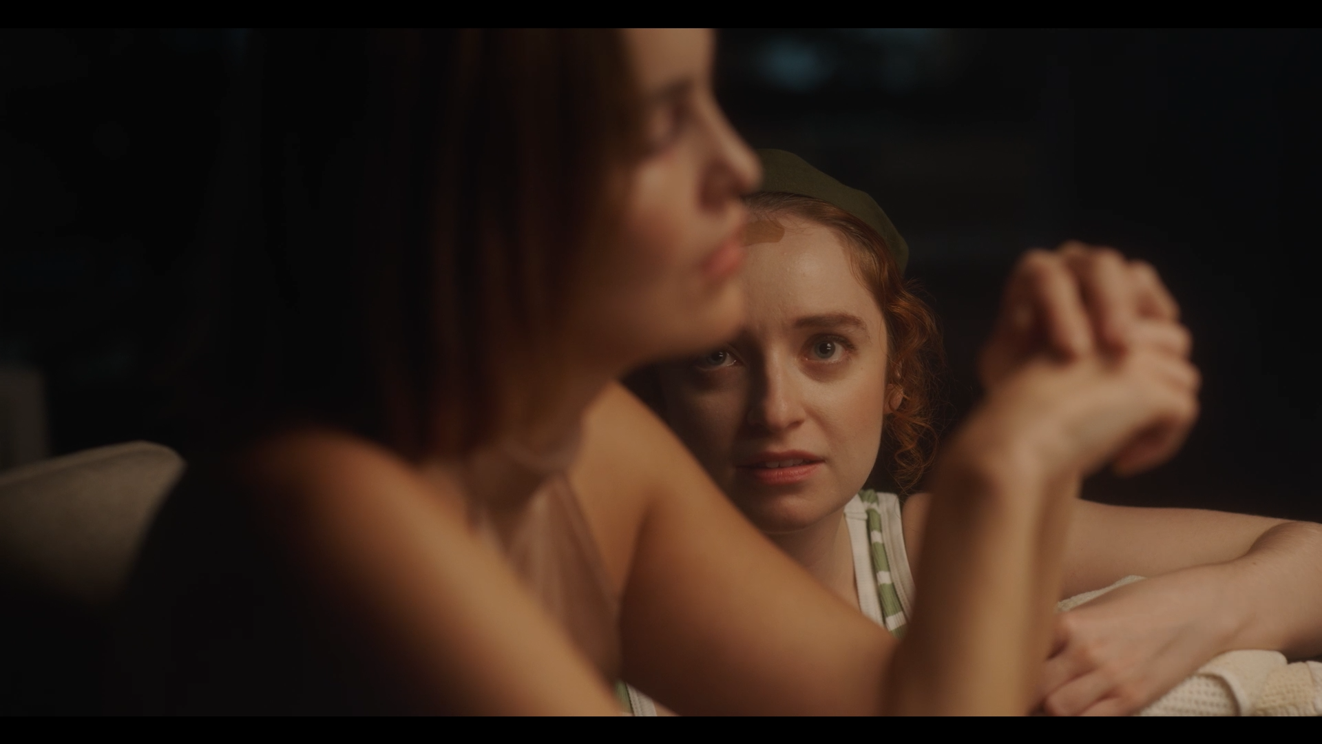 Two women sit at a table, with the woman in the background looking directly at the camera with a serious expression, while the woman in the foreground is blurred, and only part of her face is visible.