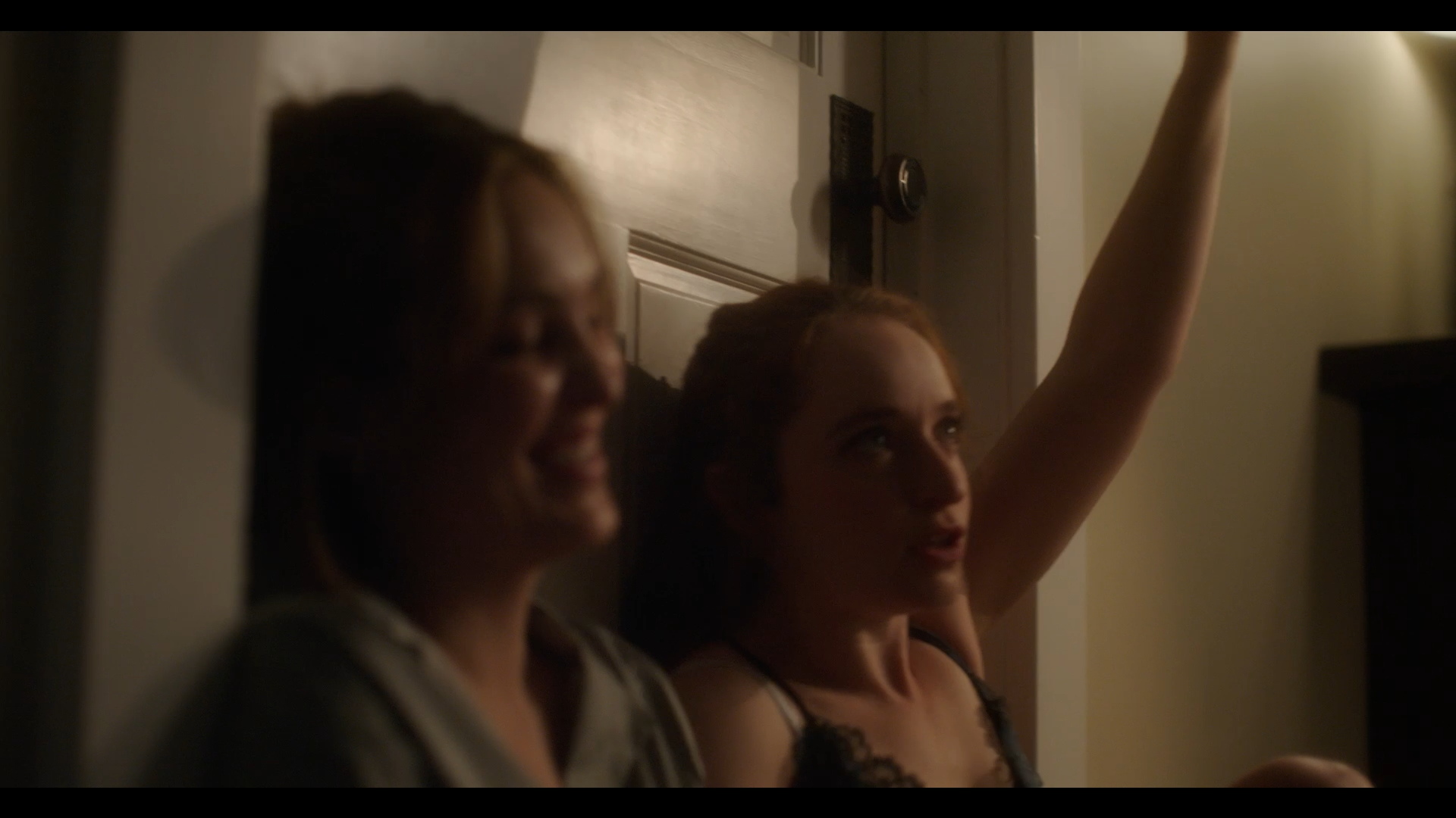 Two women sitting against a door wall, the woman on the right has her arm raised and appears to be making a point, with a background of beige walls and a door.