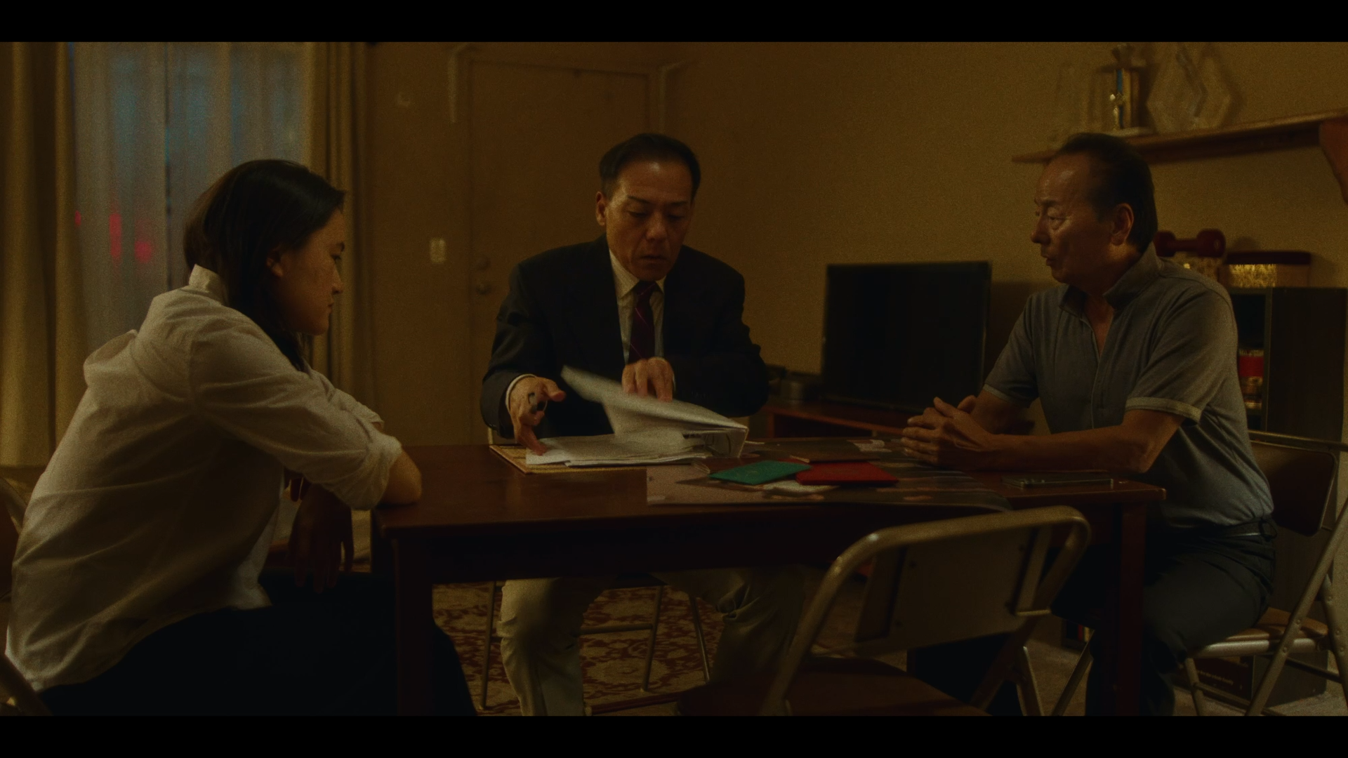 Three people sitting around a table in a dimly lit room, engaging in a discussion with documents and folders.