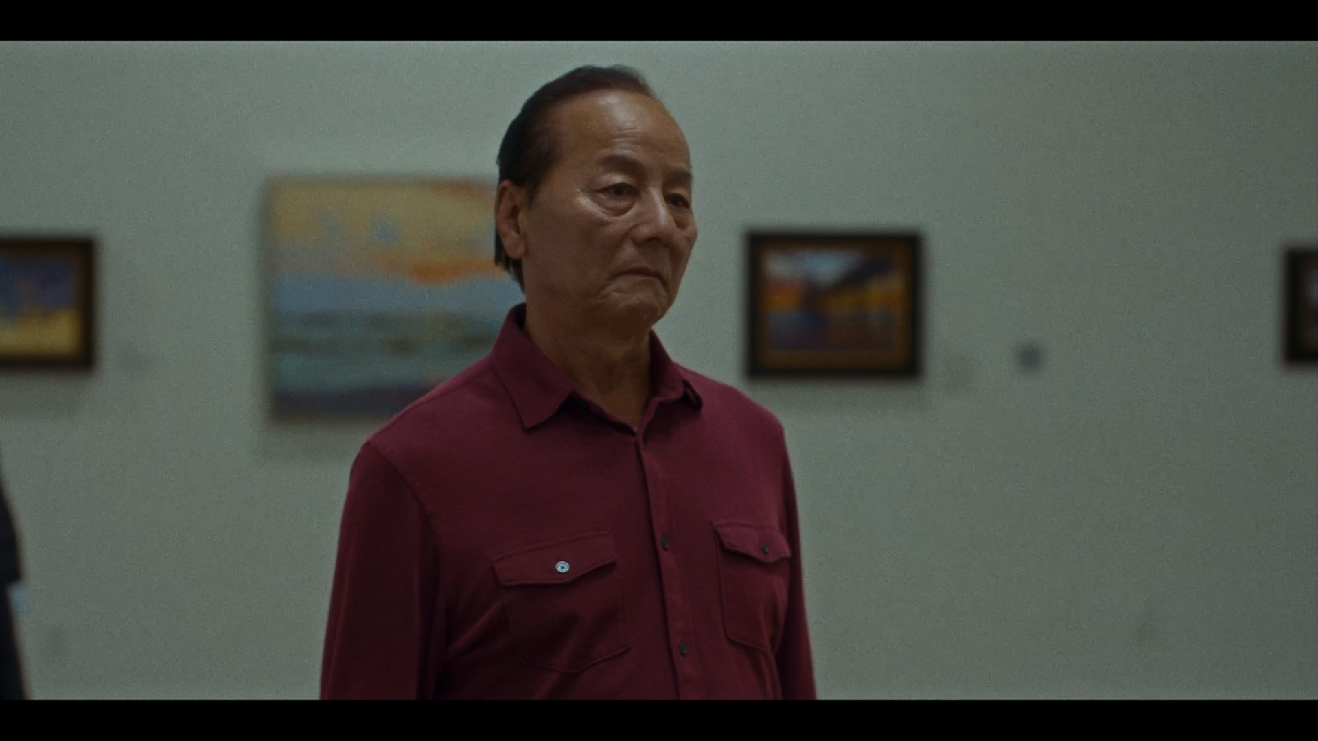 A man with slicked-back hair wearing a burgundy button-up shirt stands in front of a plain light-colored wall with framed paintings or photographs hanging on it.
