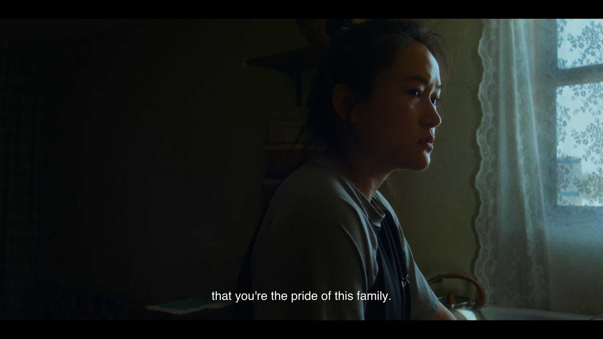 A woman sitting by a window with lace curtains, looking contemplative, with subtitles reading 'that you're the pride of this family.'