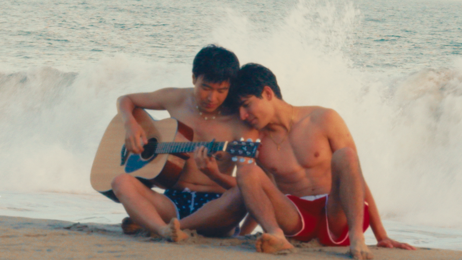 Two shirtless young men sitting on the beach at the water's edge, one playing a guitar and the other looking at a mobile phone, with waves crashing behind them.
